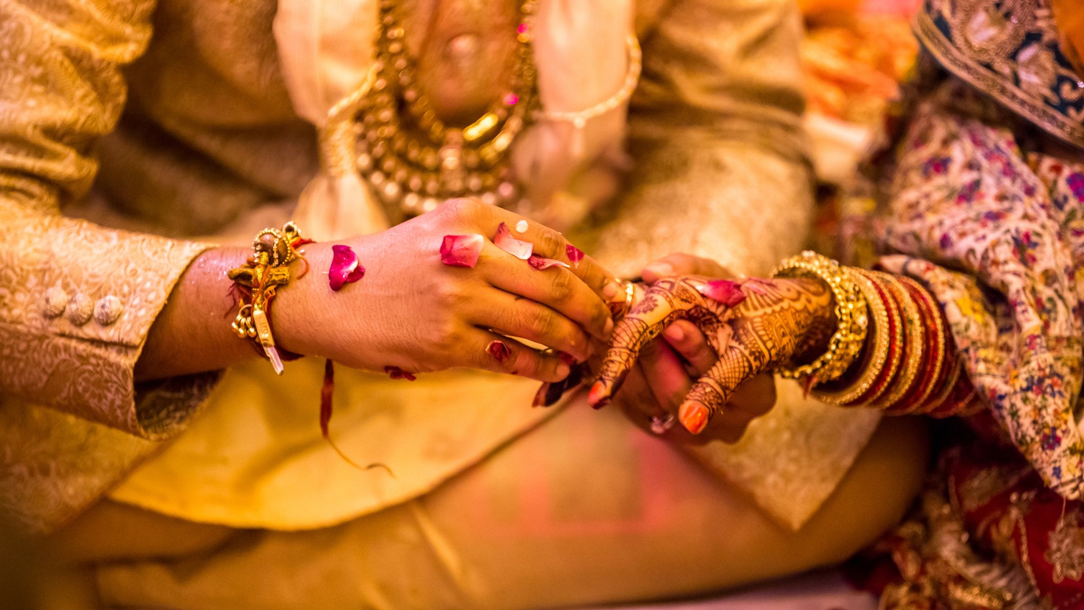 Groom placing a ring on the bride’s henna-decorated hand during a traditional wedding ceremony, with rose petals and ornate jewelry.