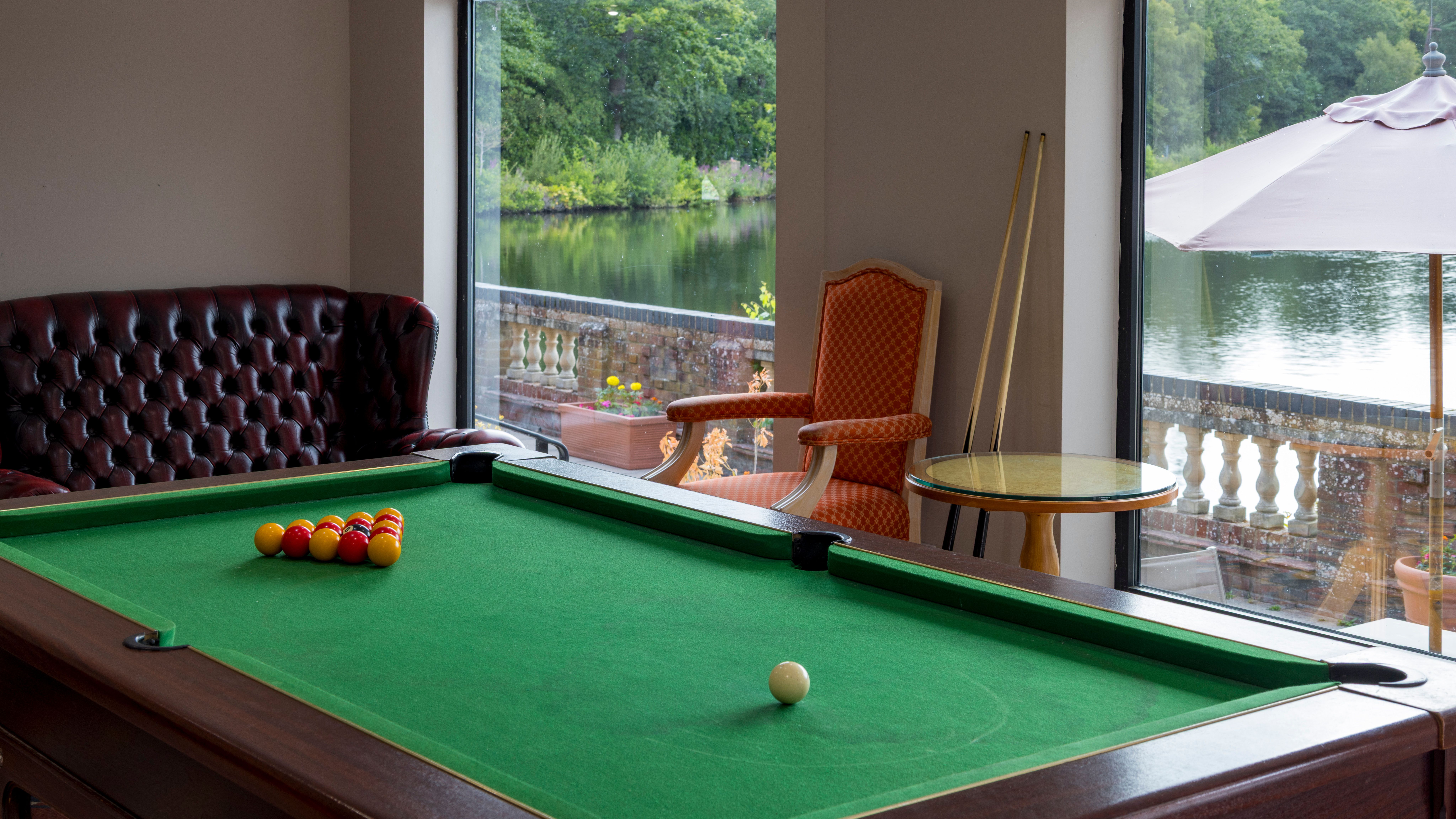 A room with a pool table, leather sofa, armchair, and large windows overlooking a lake with greenery outside.