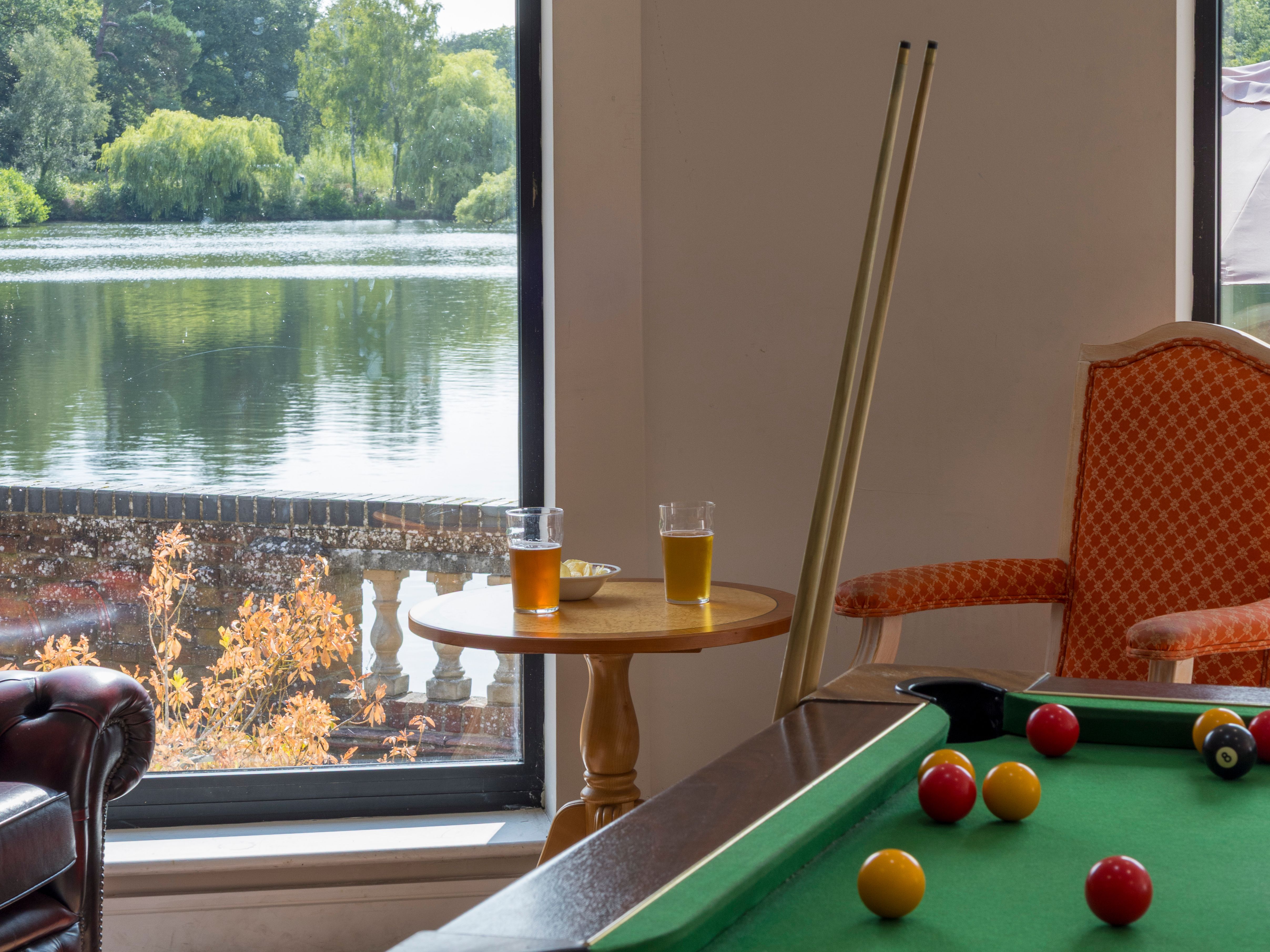 A cozy room with a pool table, drinks on a table, and large windows overlooking a lake and trees.