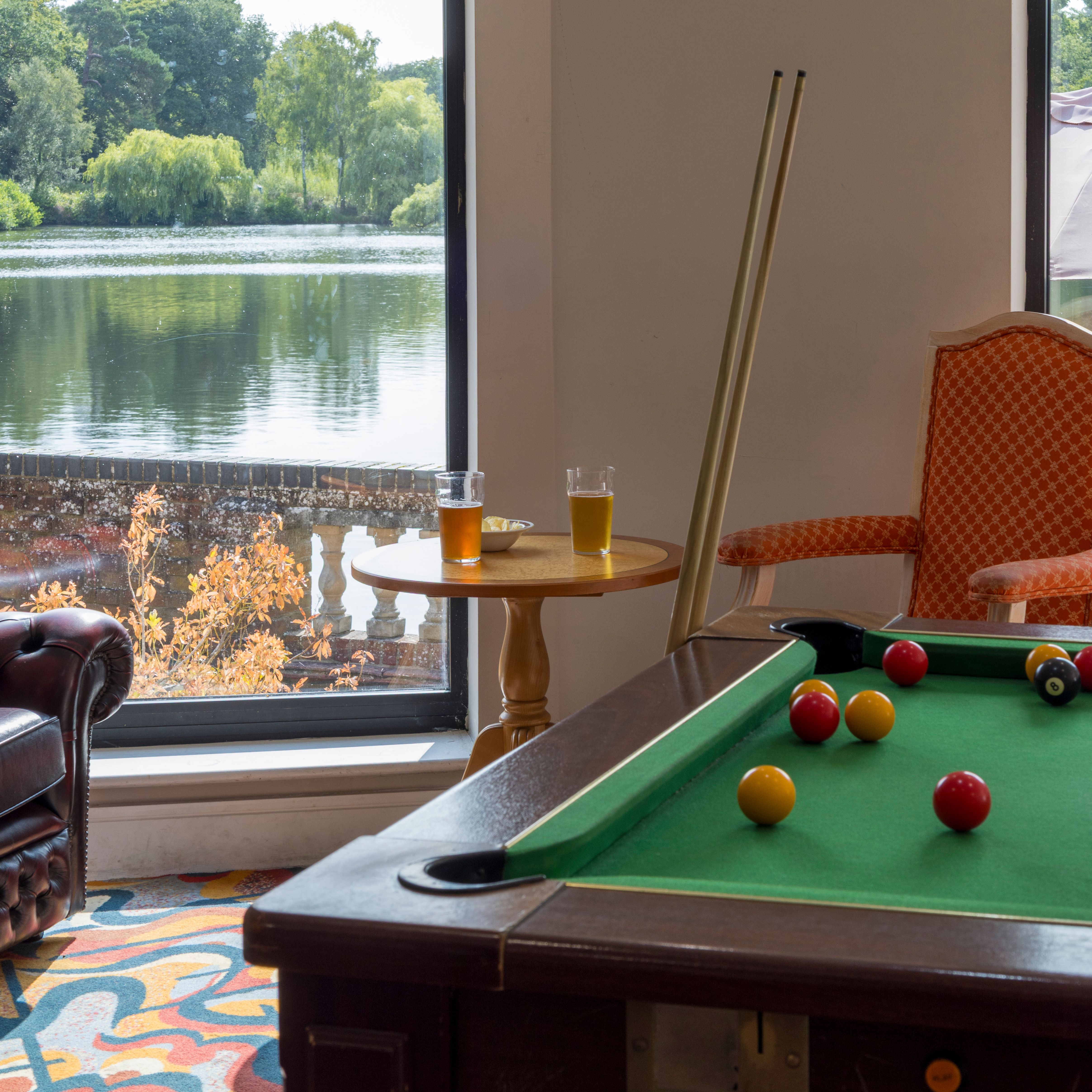 A cozy room with a pool table, drinks on a table, and large windows overlooking a lake and trees.
