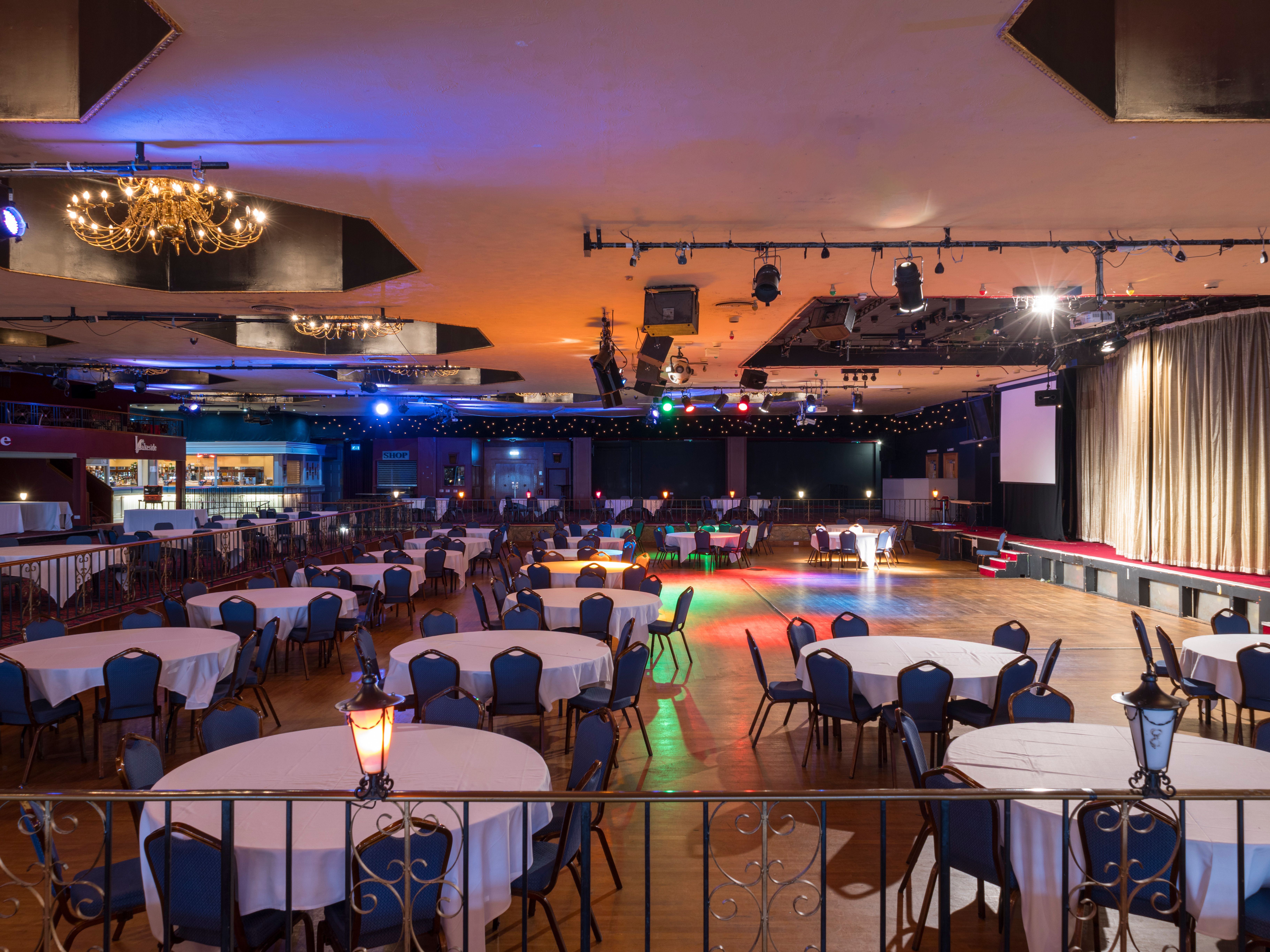Large banquet hall with round tables, blue chairs, chandeliers, and a stage with gold curtains.