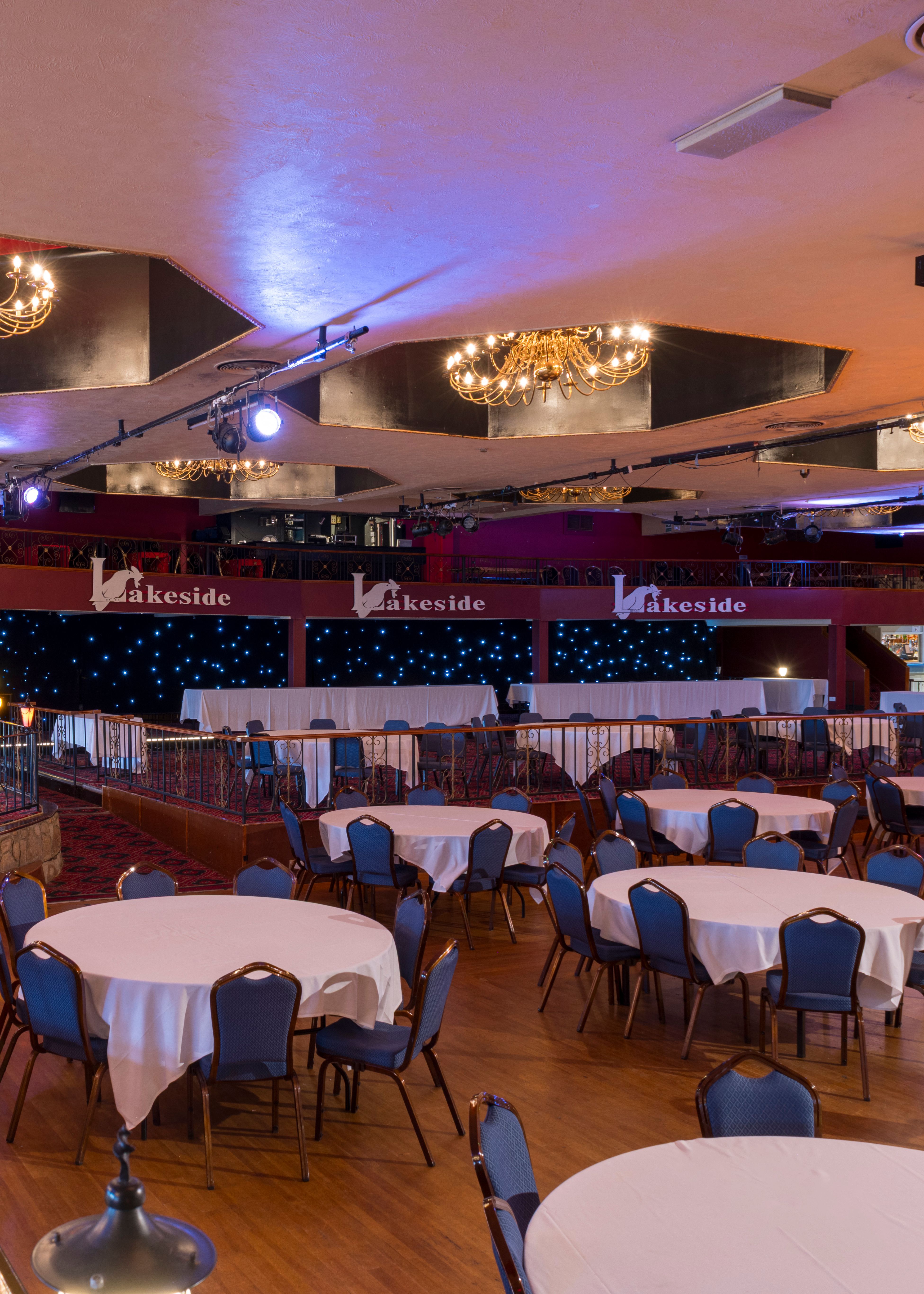 Interior of a spacious banquet hall with round tables covered in white cloths and blue chairs, chandeliers overhead, and a stage at the front.