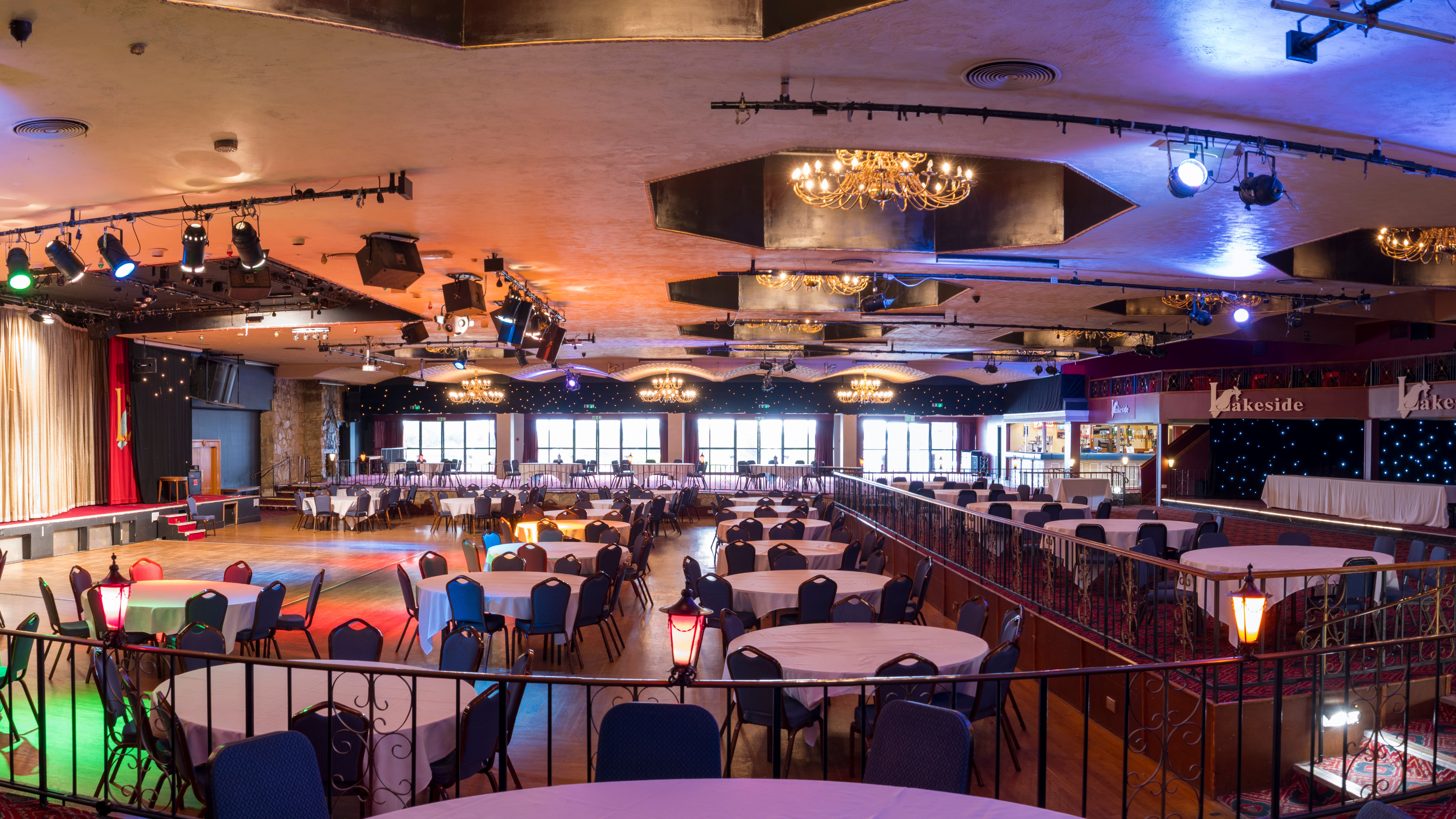Interior of a large, elegant banquet hall with round tables and chairs, stage, chandeliers, and decorative lighting