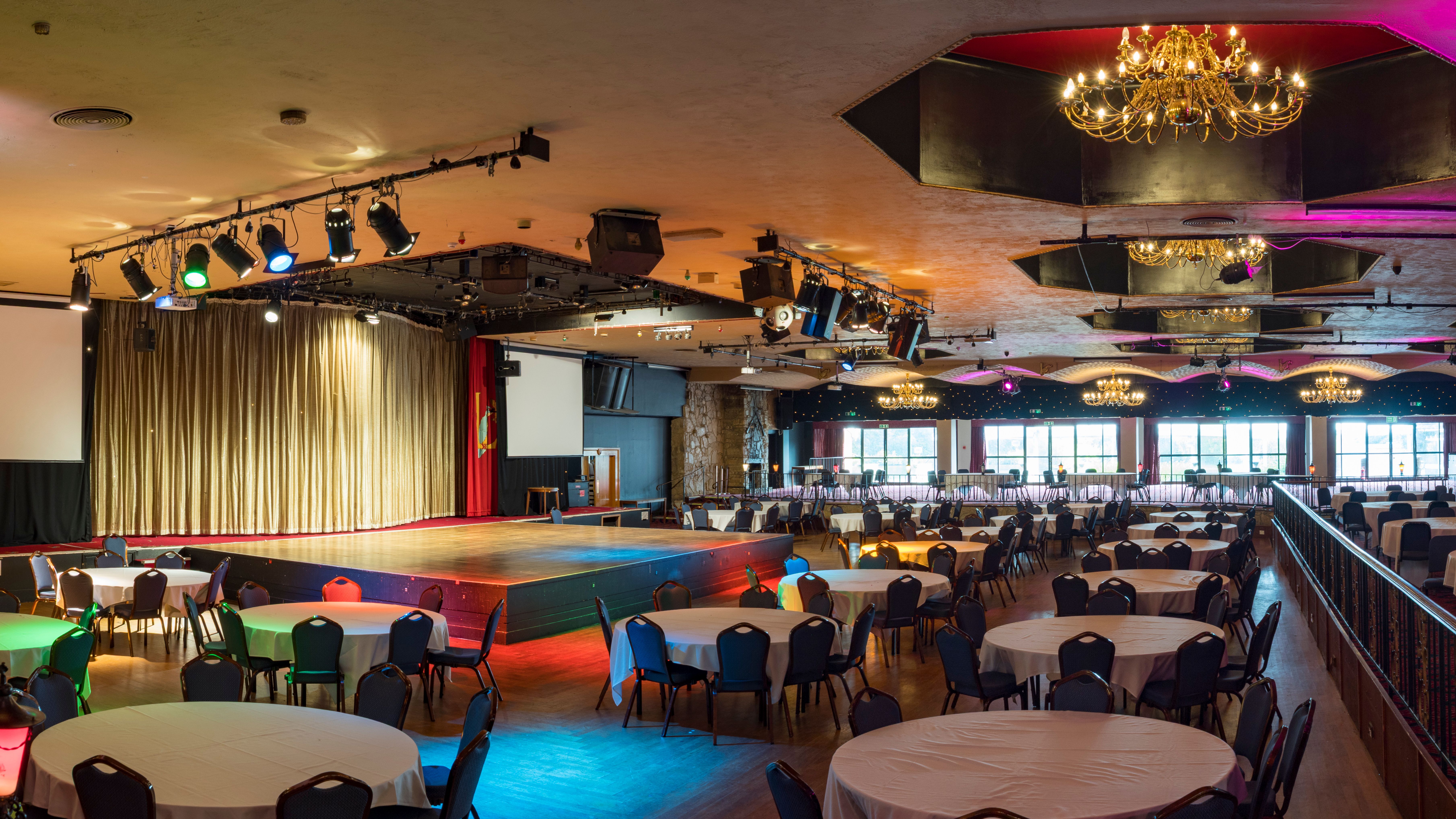 Large banquet hall with round tables, stage, chandeliers, and colorful lighting