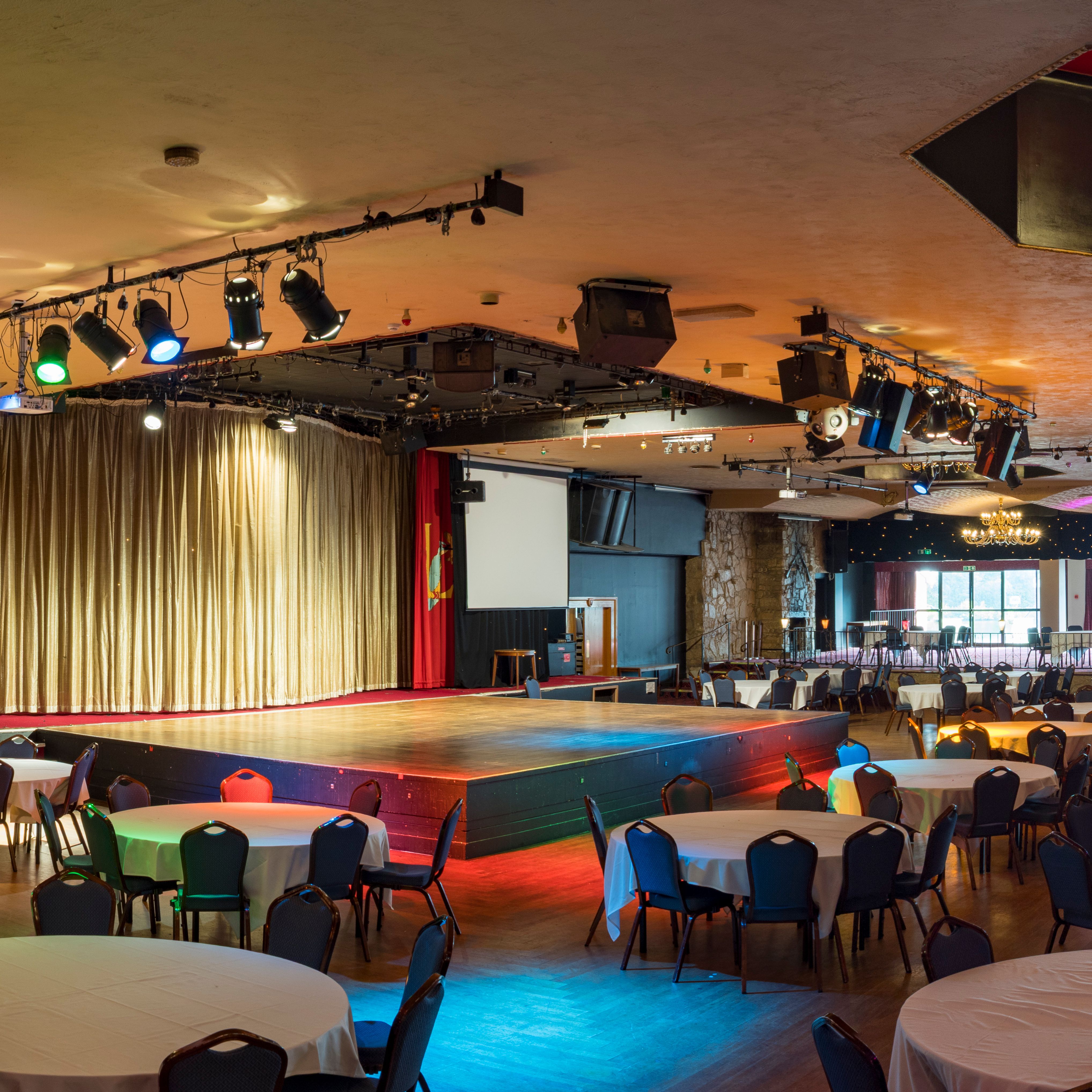 Large banquet hall with round tables, stage, chandeliers, and colorful lighting