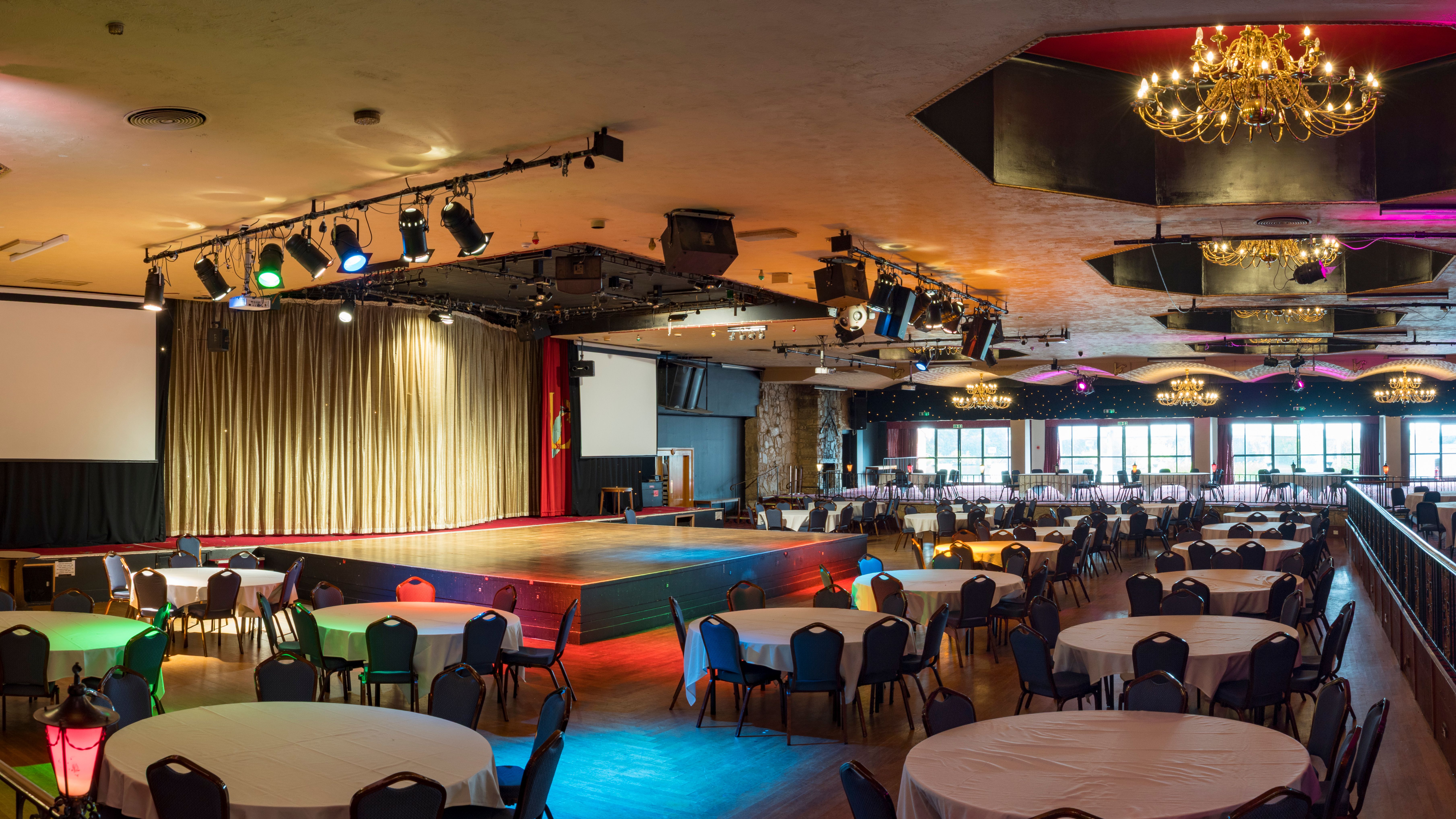 Large banquet hall with round tables, stage, chandeliers, and colorful lighting