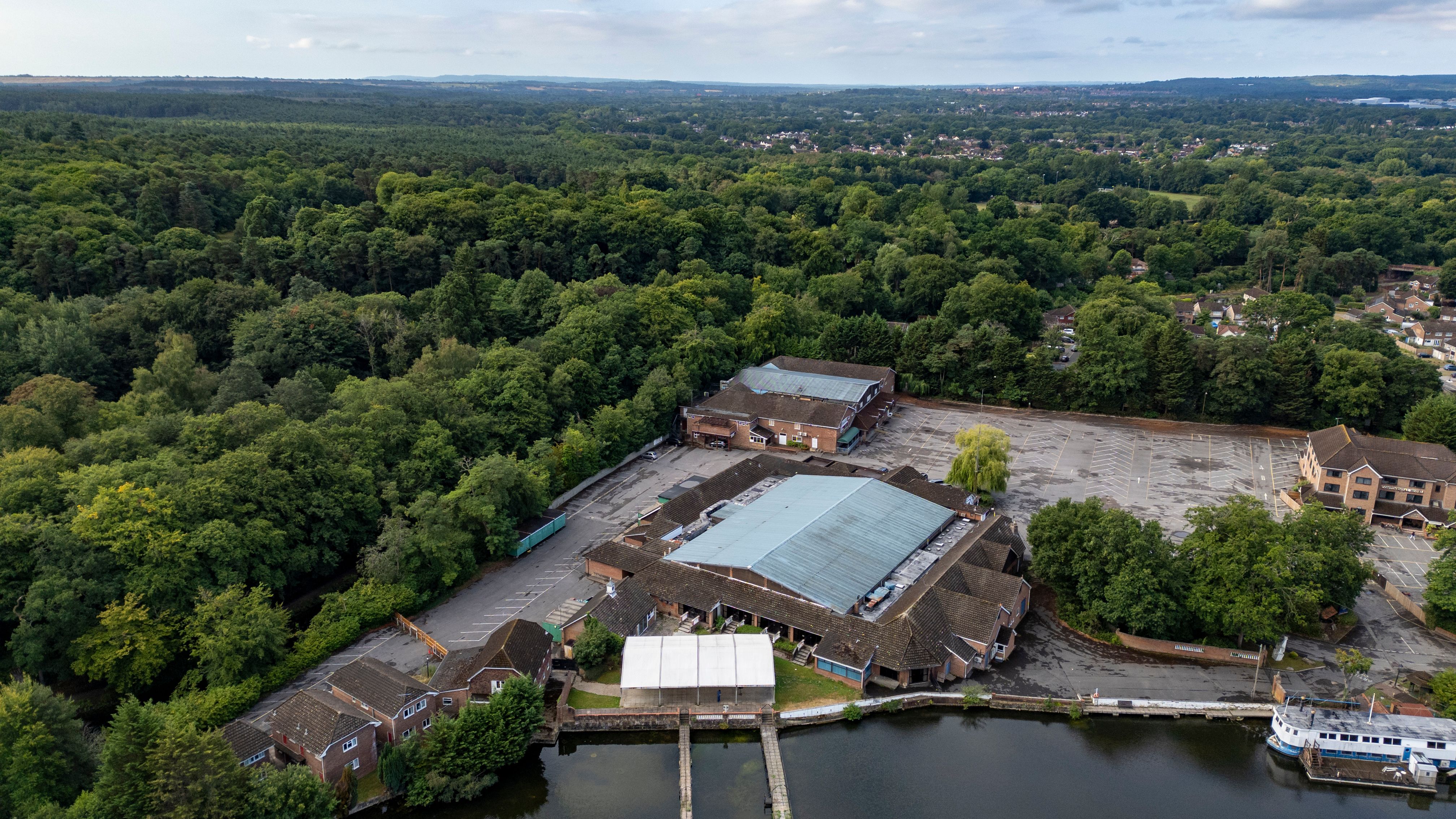 Aerial view of a large building complex near a lake, surrounded by dense forest and a few scattered houses.