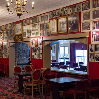 Interior of a room with red walls covered in framed photographs, tables and chairs, and chandeliers hanging from the ceiling.