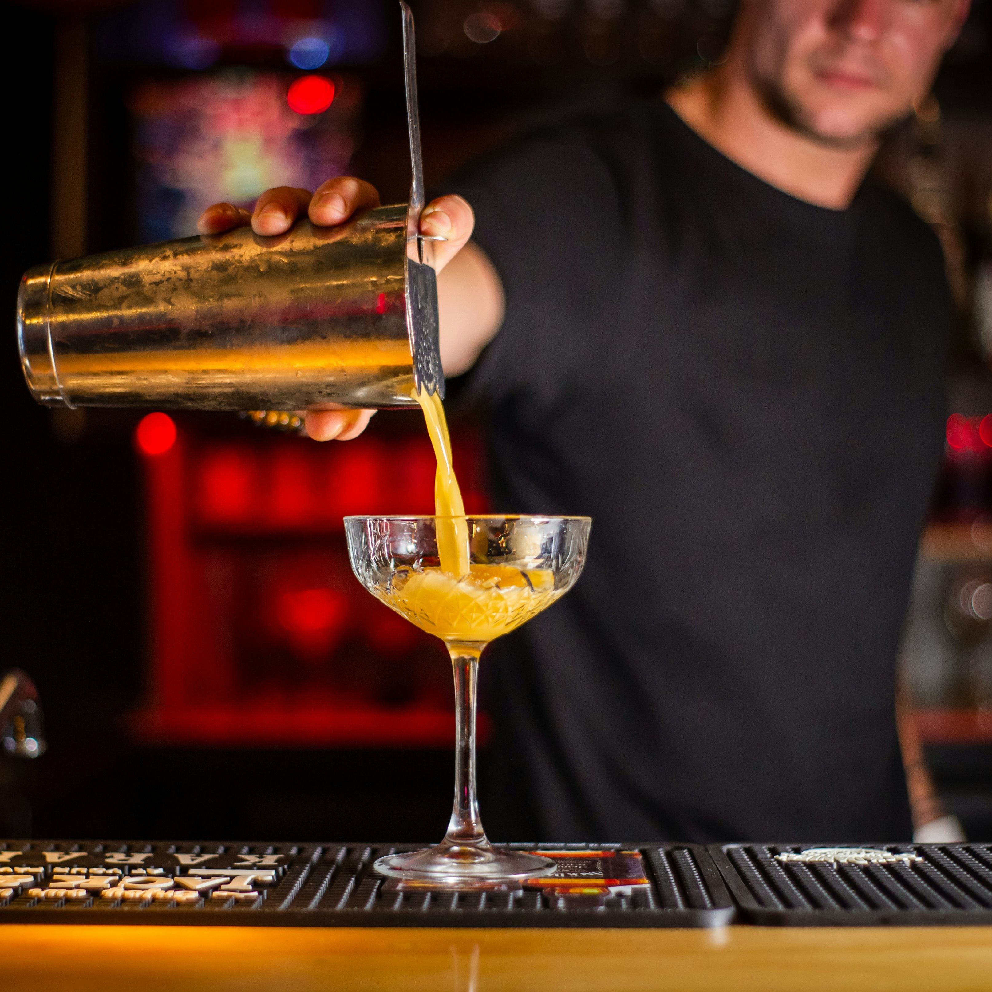 Bartender pouring a yellow cocktail from a shaker into a glass at a bar.