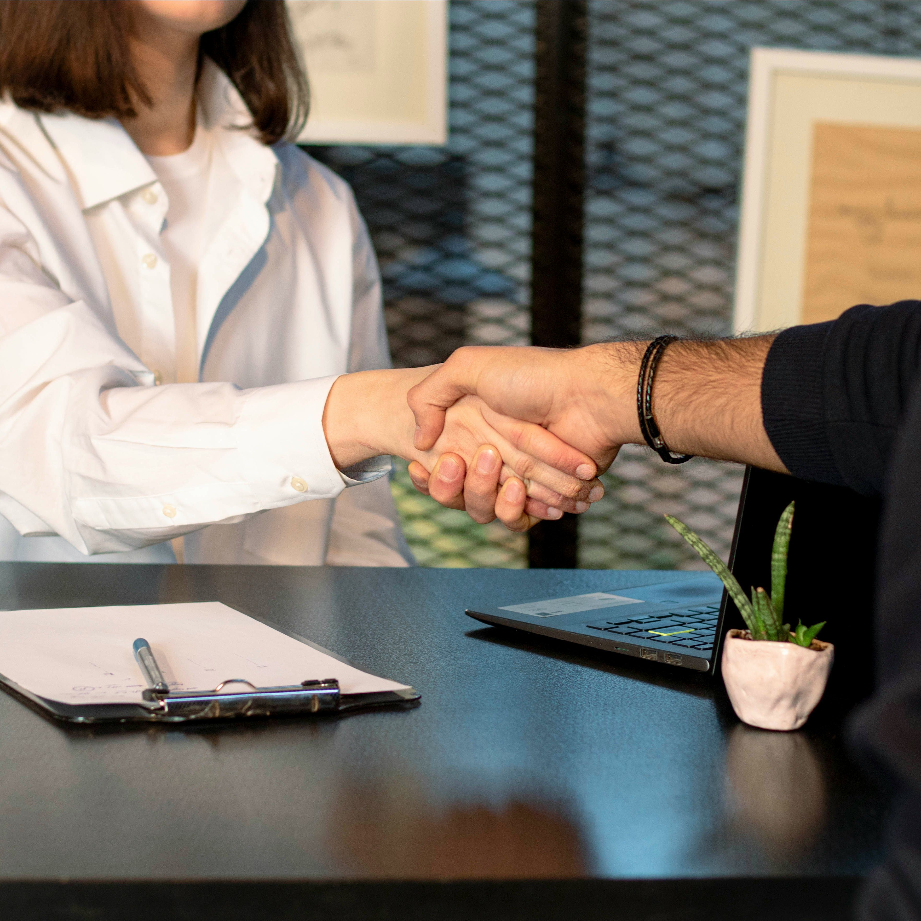 Two people shaking hands at a desk with a clipboard, laptop, and small potted plant