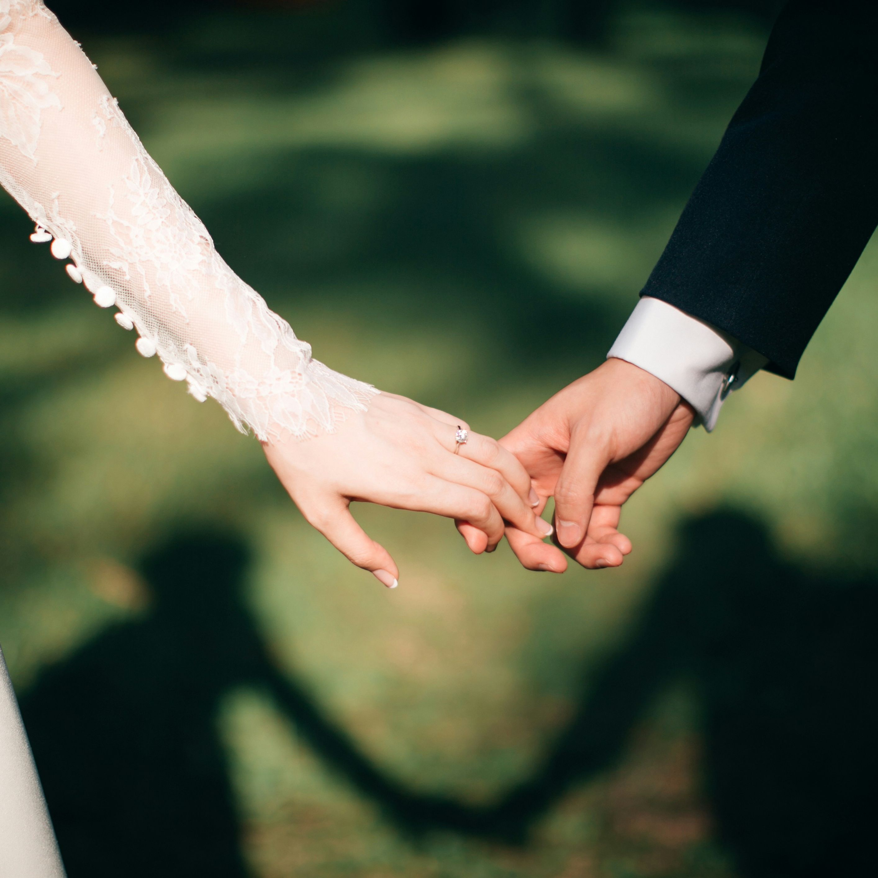 Close-up of a bride and groom holding hands outdoors with their shadows in the background.