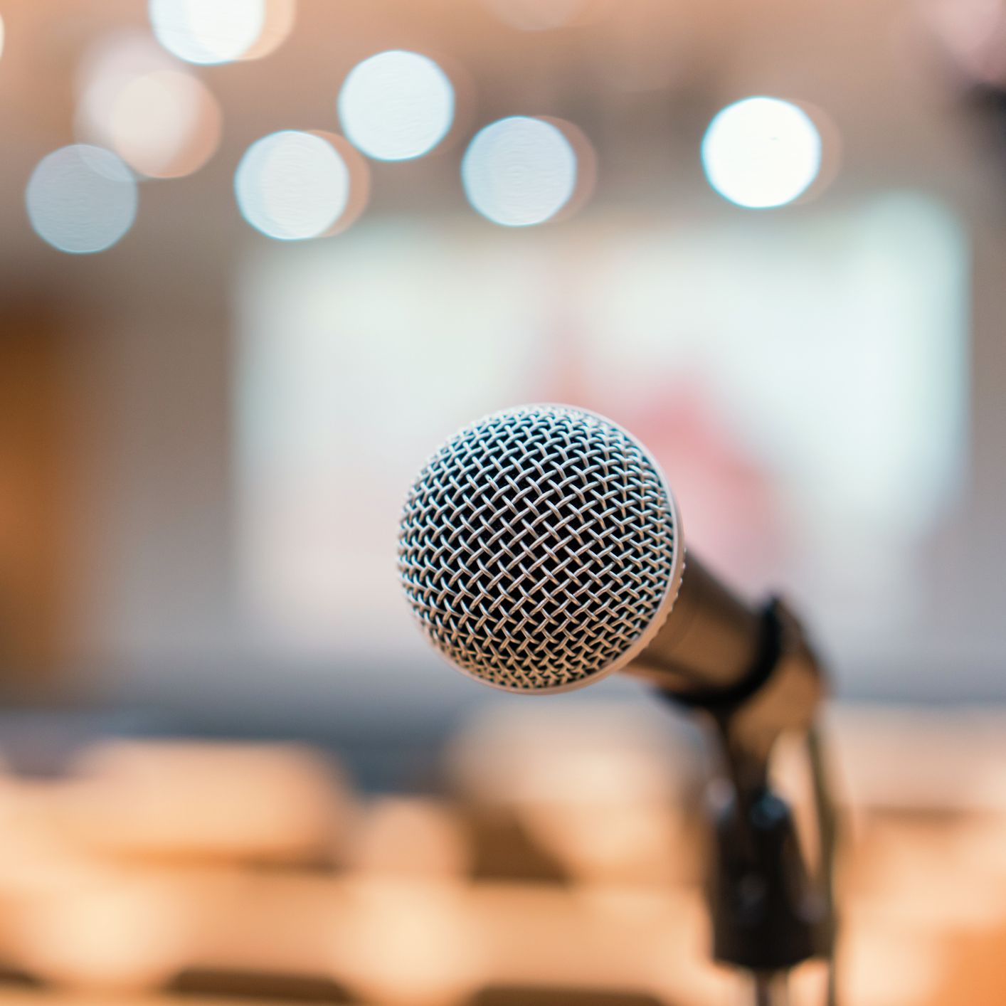 Close-up of a microphone on a stand with a blurred background of lights and empty seats, suggesting a stage or conference setting.
