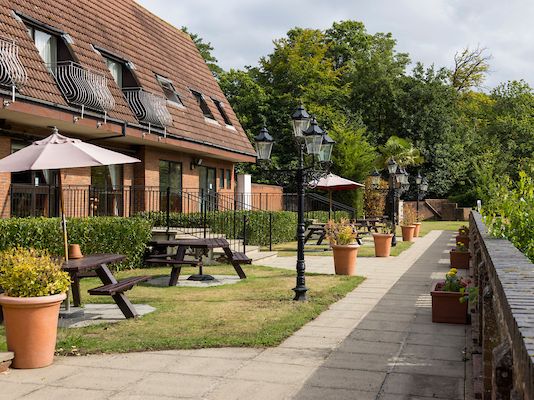 Outdoor seating area with picnic tables, umbrellas, and potted plants next to a brick building and walkway.