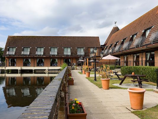 Hotel or lodge with red tiled roofs next to a calm lake, outdoor seating and potted plants along a paved walkway.