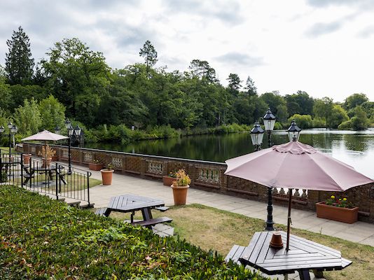 Lakeside outdoor seating area with picnic tables, umbrellas, and a view of a calm lake surrounded by trees.