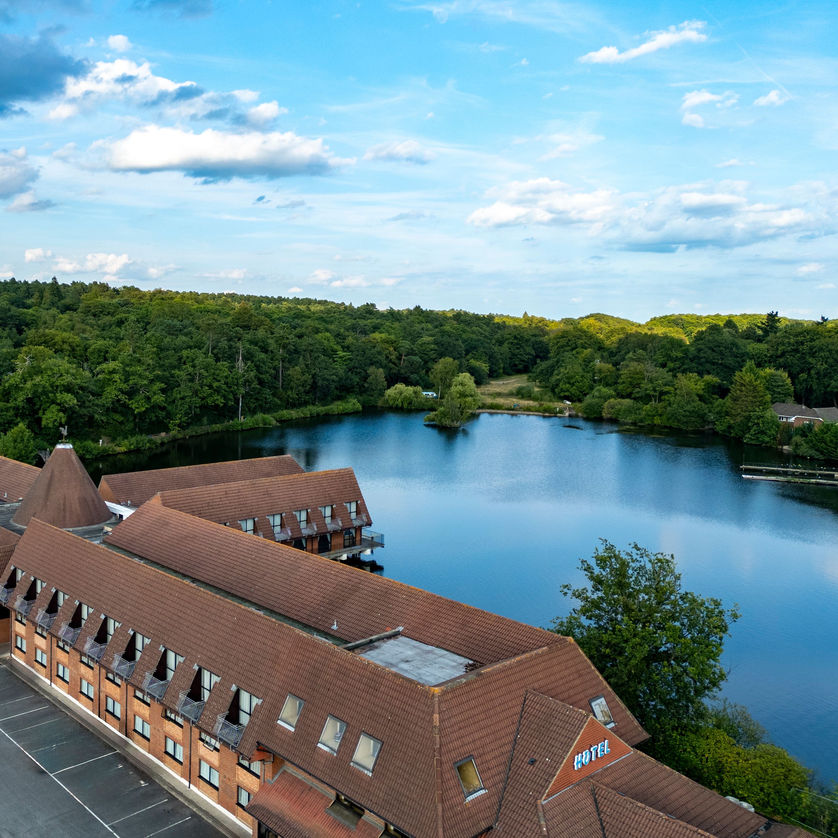 Aerial view of a hotel building beside a lake surrounded by lush green forest under a blue sky with clouds.