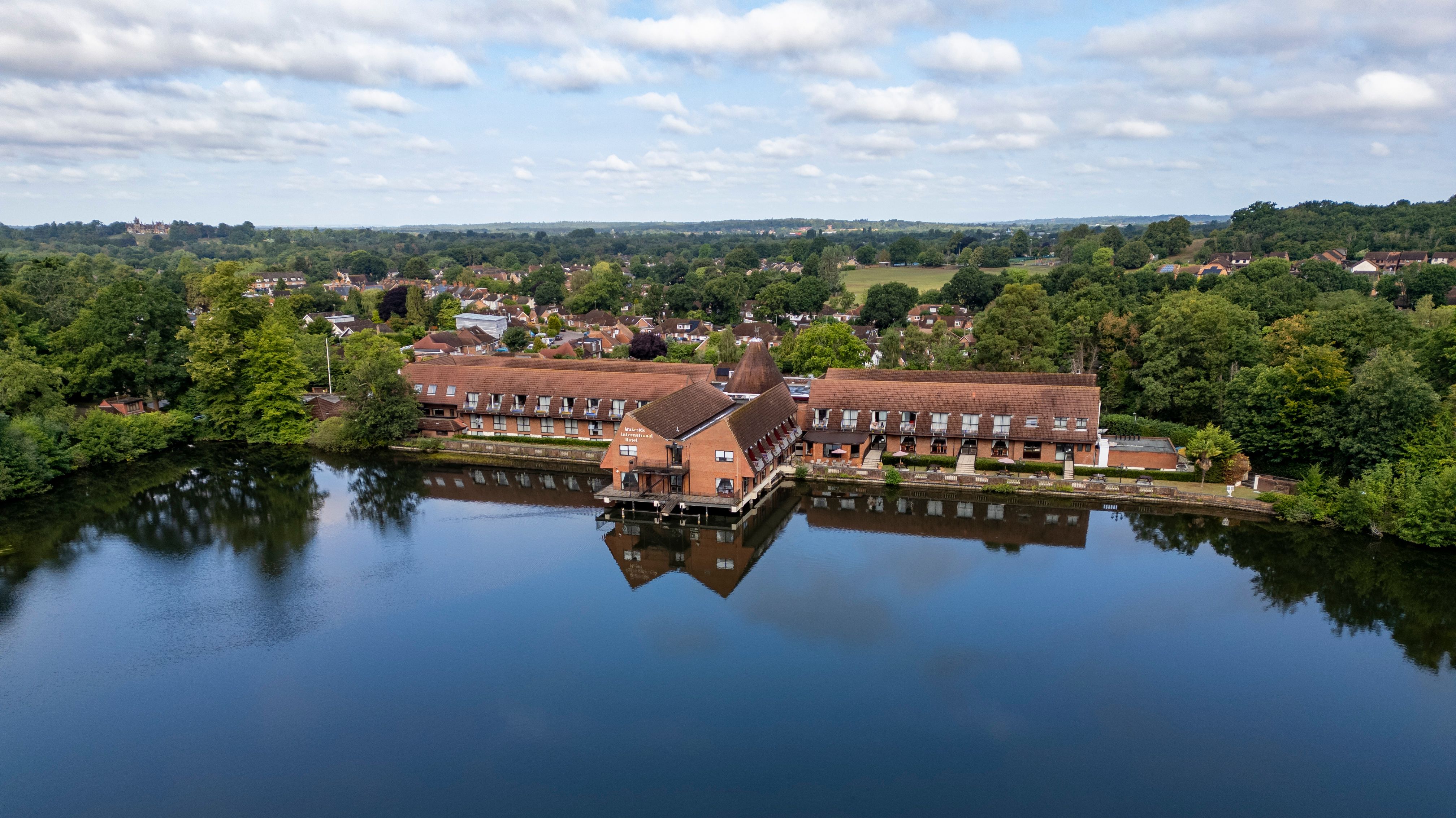 Aerial view of a large brick building complex situated on the edge of a calm lake, surrounded by greenery and trees.
