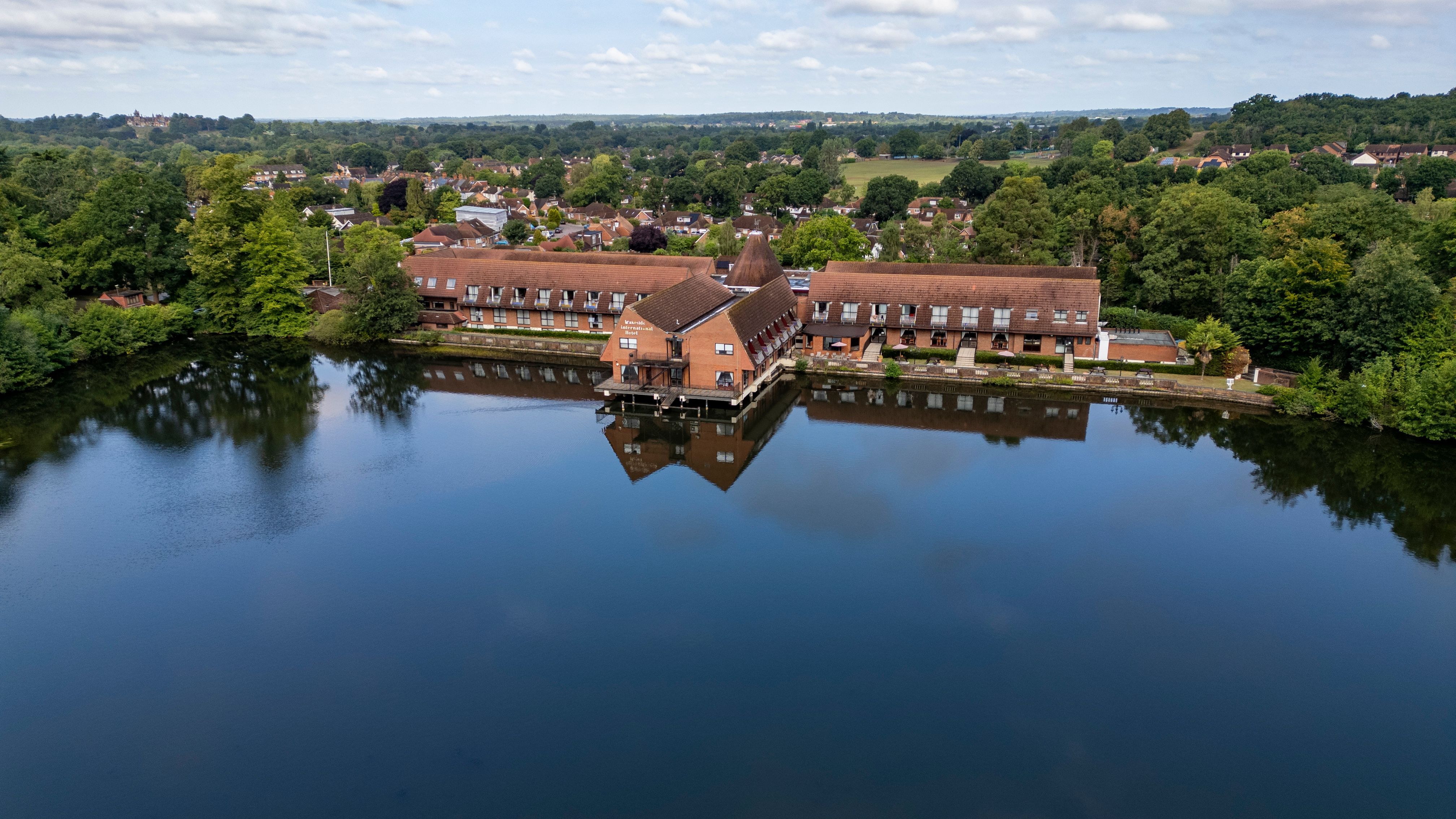 Aerial view of a large brick building complex situated on the edge of a calm lake, surrounded by greenery and trees.
