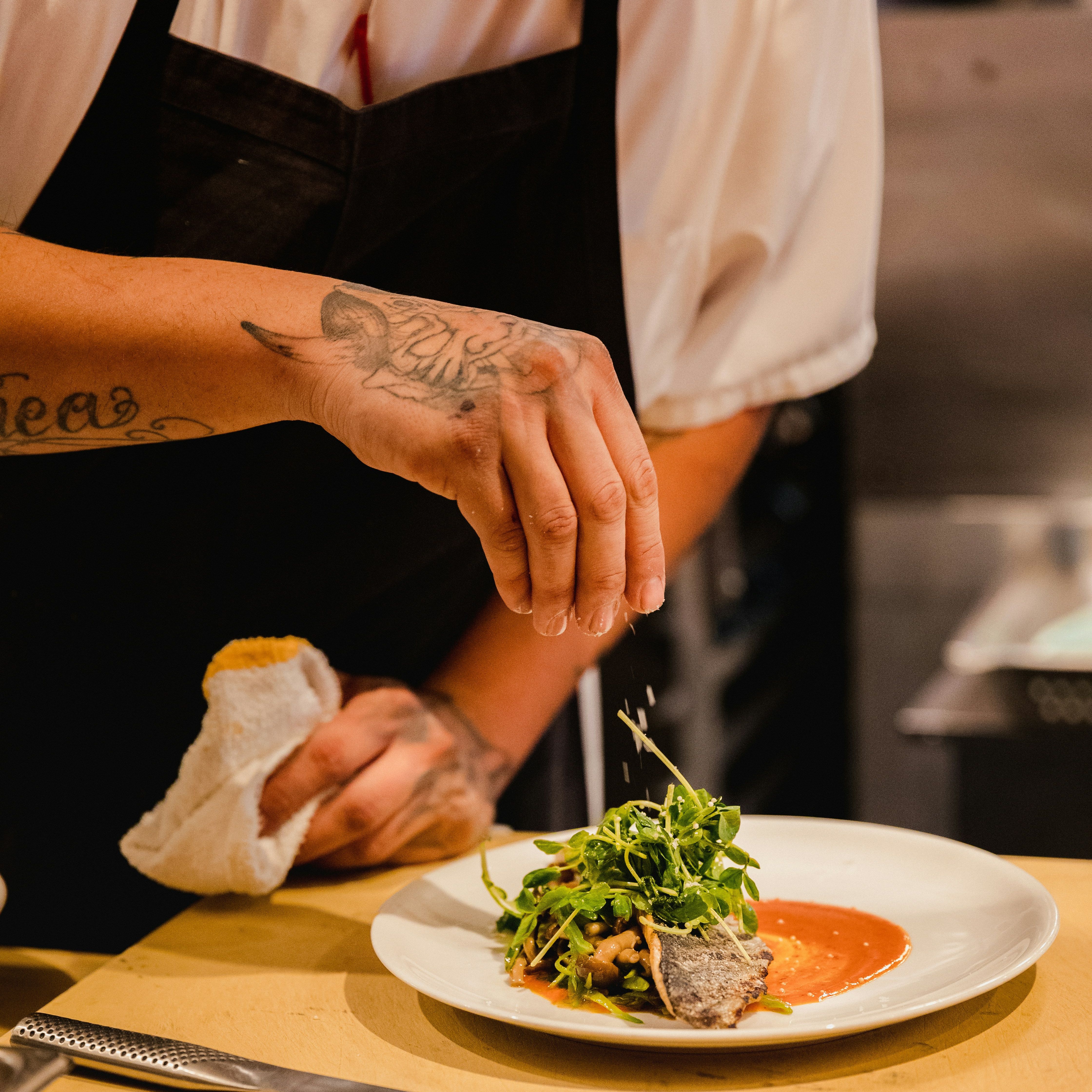 Chef garnishing a plated dish with fresh greens and sauce in a professional kitchen