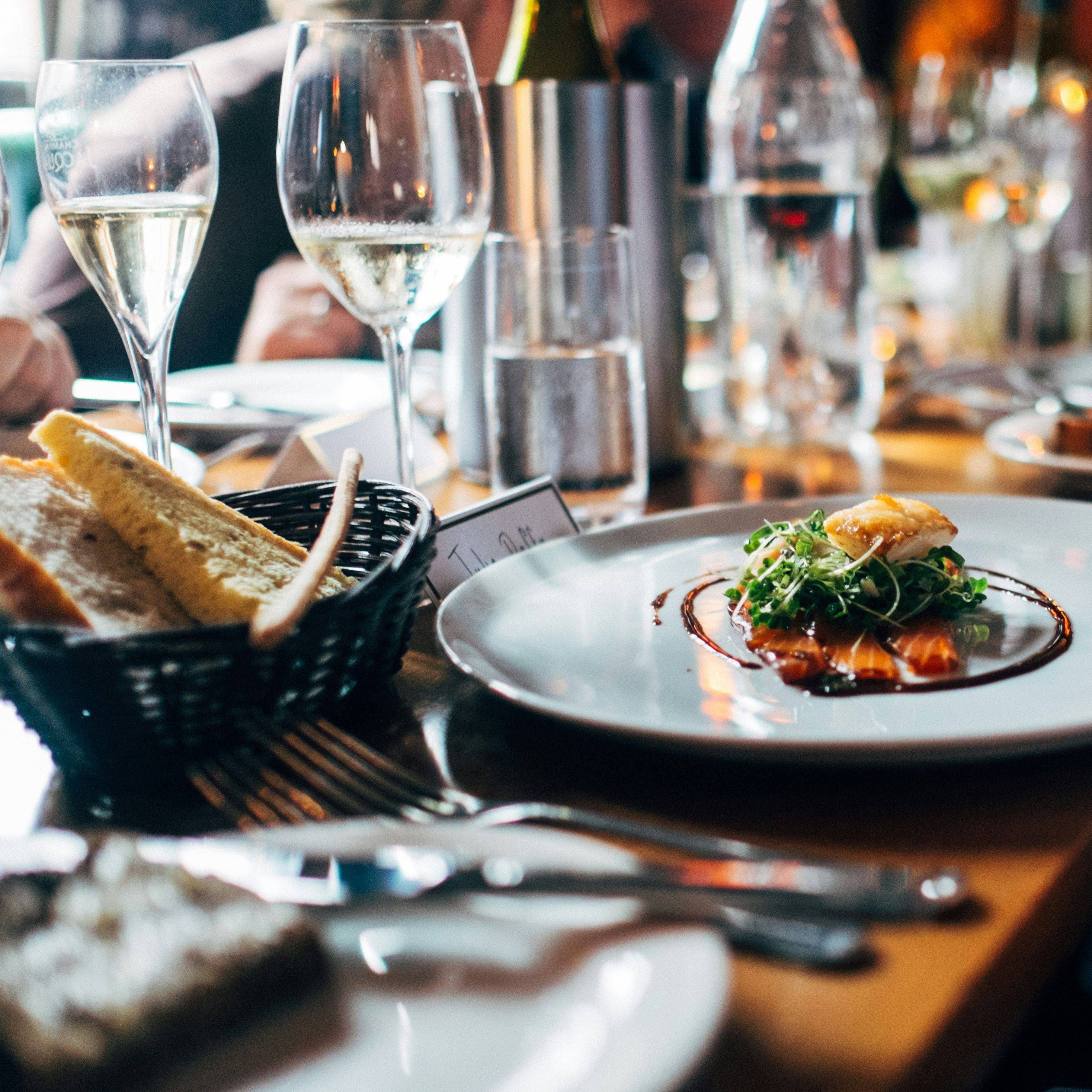 Elegant restaurant table setting with fine dining dish, bread basket, and glasses of white wine.