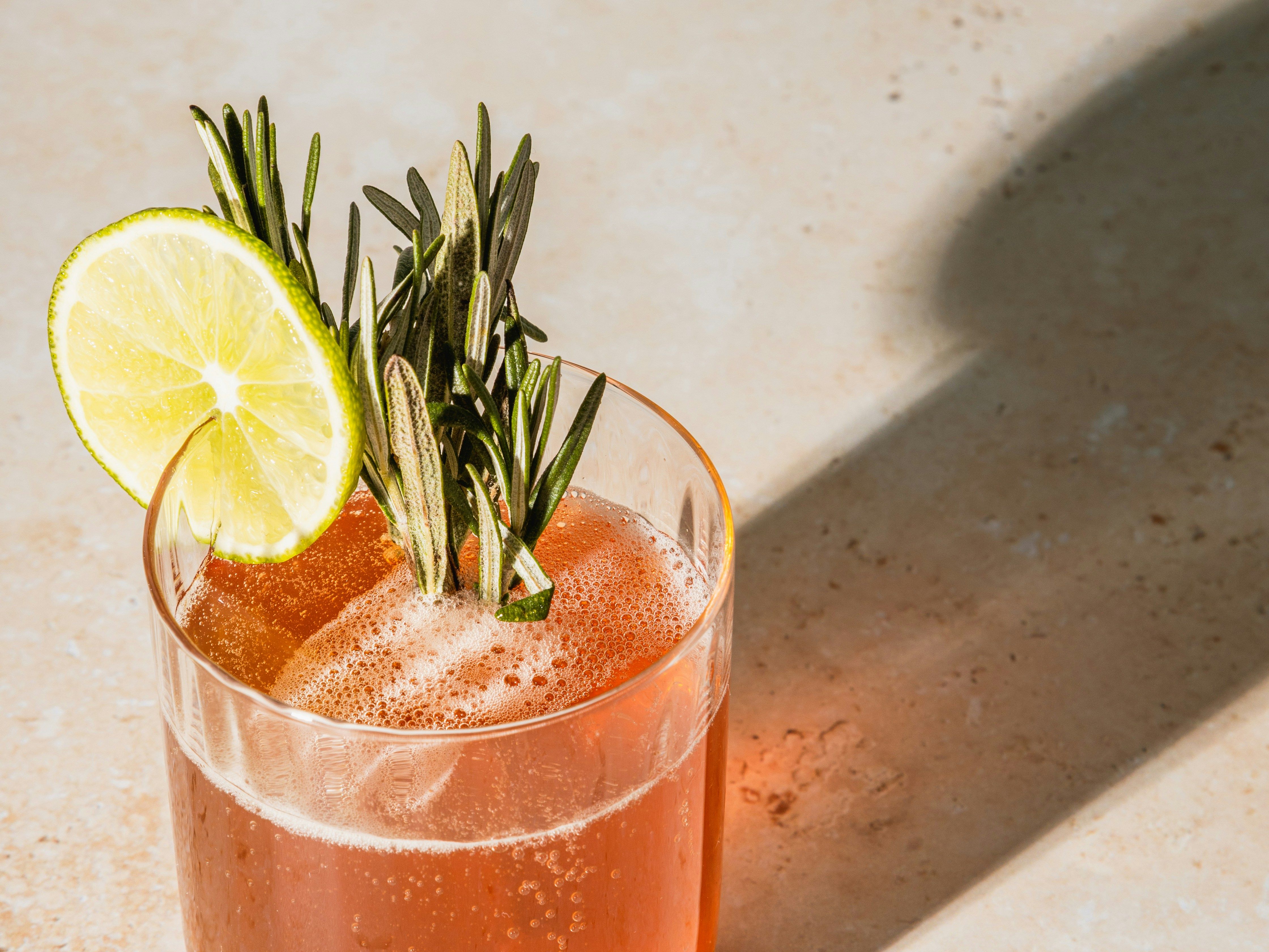 A glass of fizzy reddish cocktail garnished with a slice of lime and sprigs of rosemary.