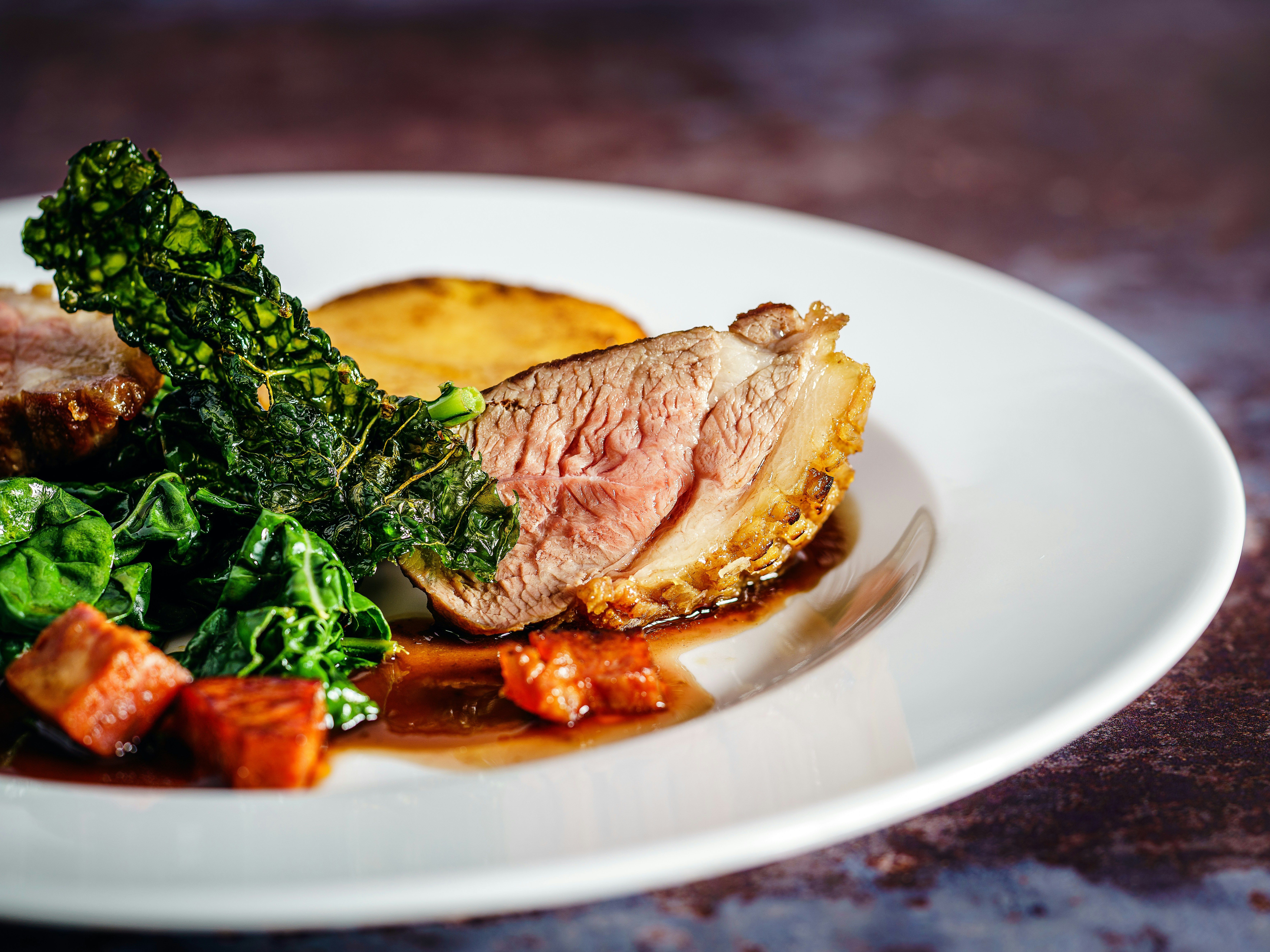 Gourmet plated dish featuring a slice of medium-cooked meat with crispy skin, leafy greens, a potato round, and sauce on a white plate.