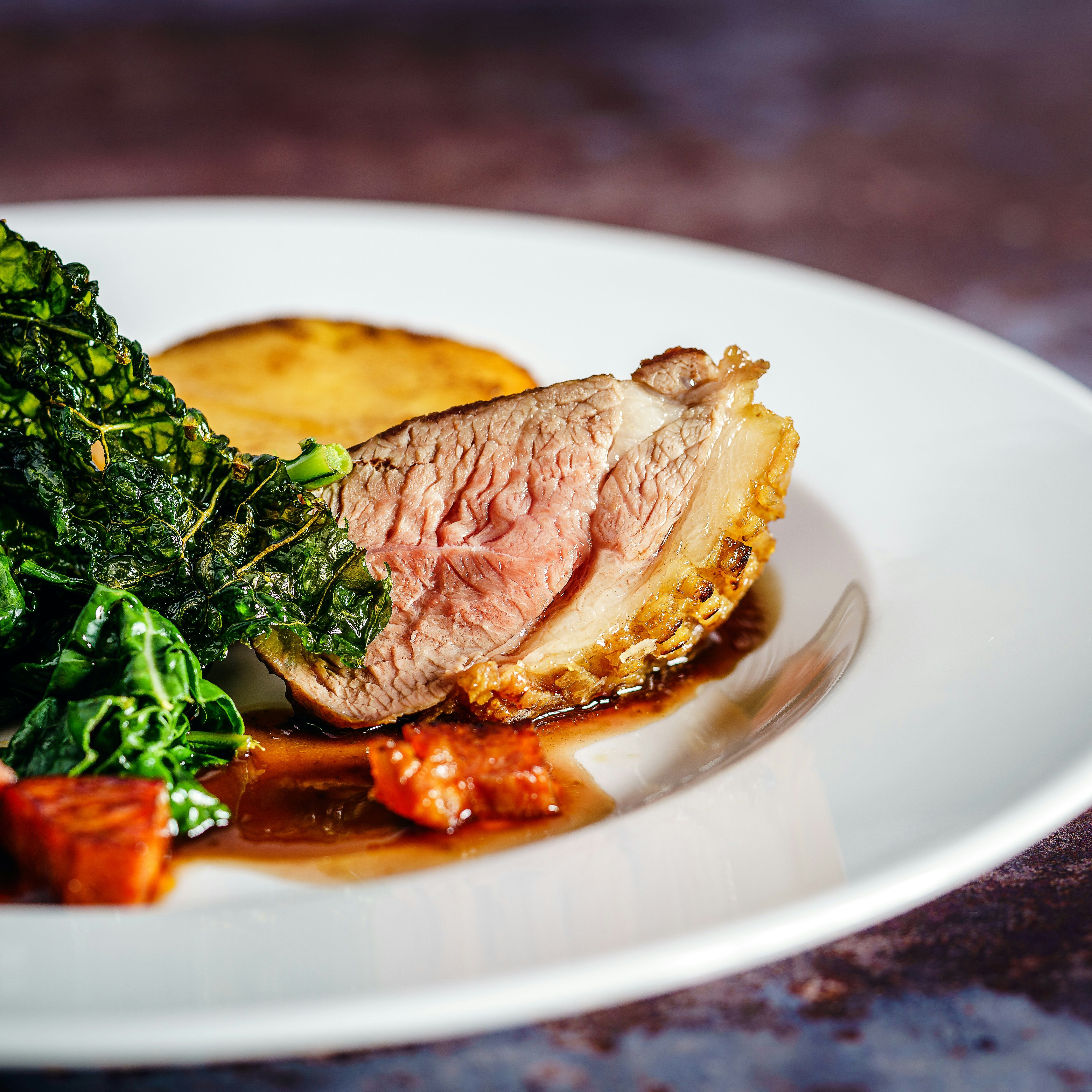 Gourmet plated dish featuring a slice of medium-cooked meat with crispy skin, leafy greens, a potato round, and sauce on a white plate.