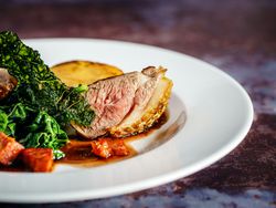 Gourmet plated dish featuring a slice of medium-cooked meat with crispy skin, leafy greens, a potato round, and sauce on a white plate.