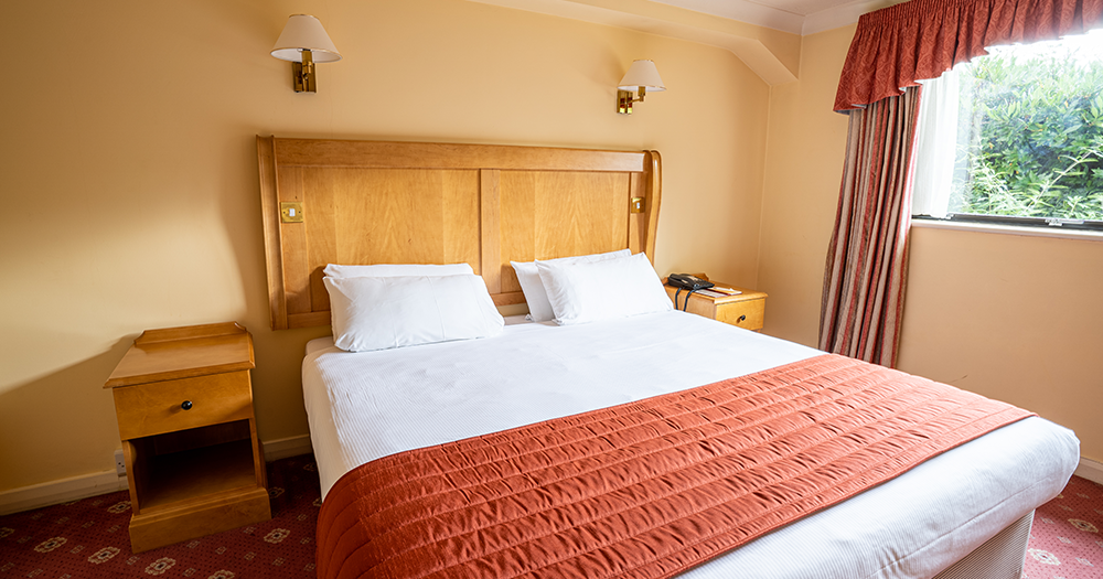 A neatly made bed with white sheets and an orange blanket in a cozy hotel room with wooden furniture.