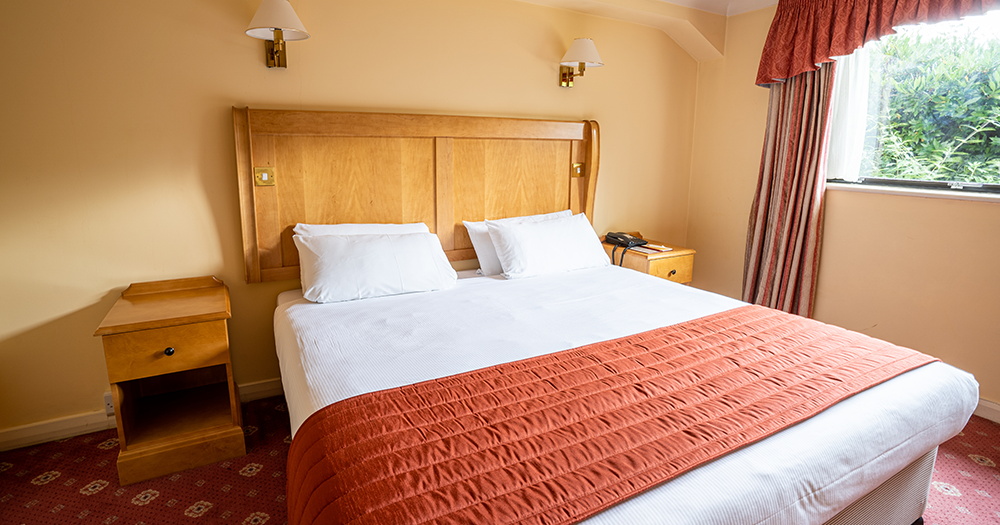 A neatly made bed with white sheets and an orange blanket in a cozy hotel room with wooden furniture.