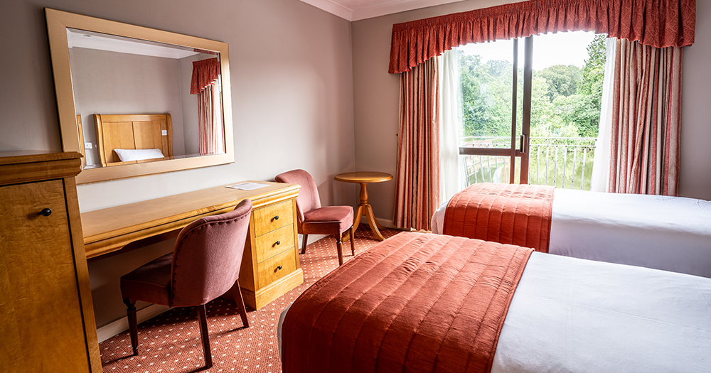 A hotel room with twin beds, a desk, chairs, and a large window with a view of greenery outside.