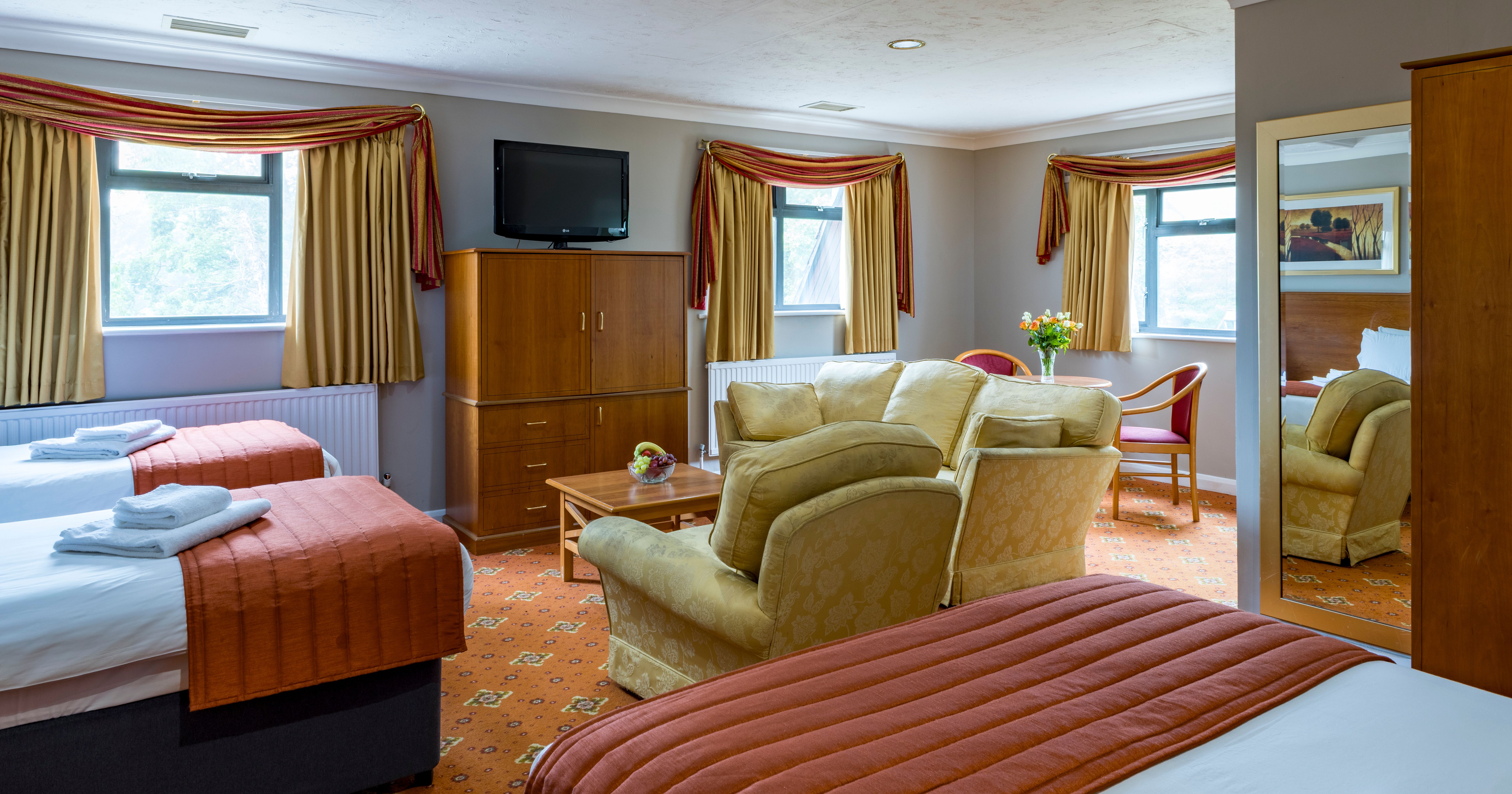 Spacious hotel room with two beds, a sitting area, and large windows with gold curtains.
