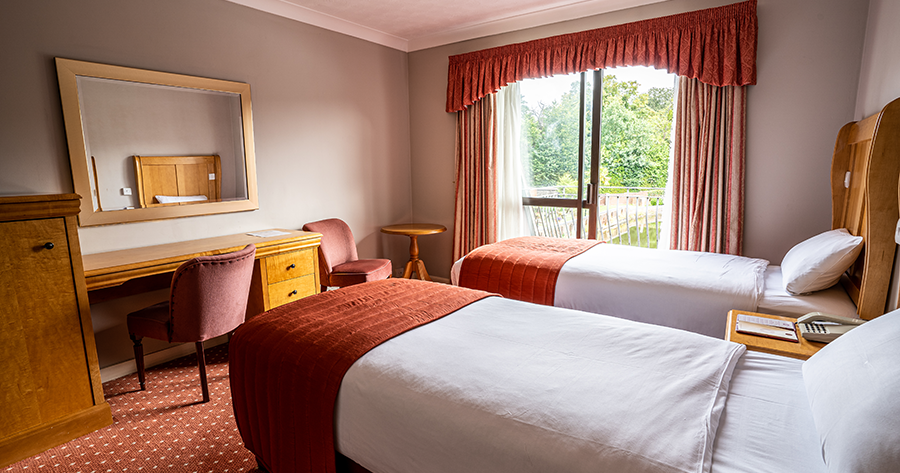 Hotel room with two single beds, a desk, mirror, and large window with curtains.