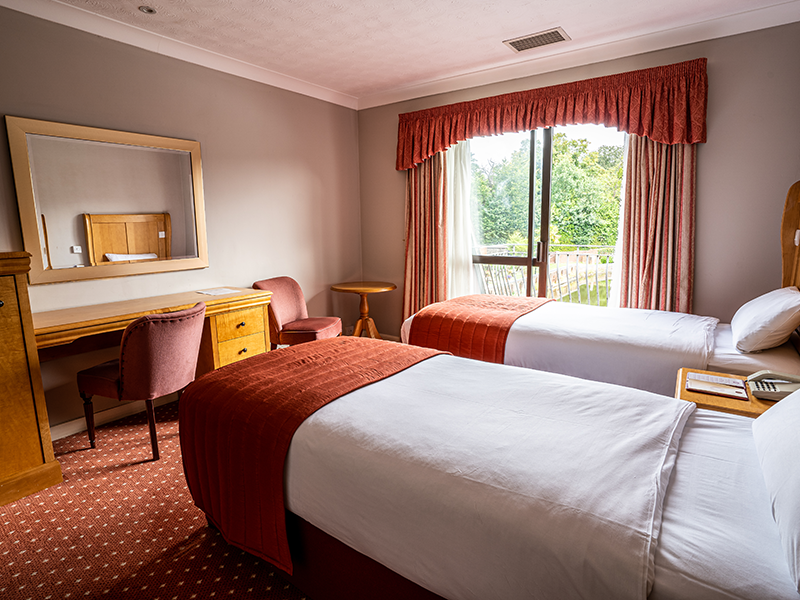 Hotel room with two single beds, a desk, mirror, and large window with curtains.