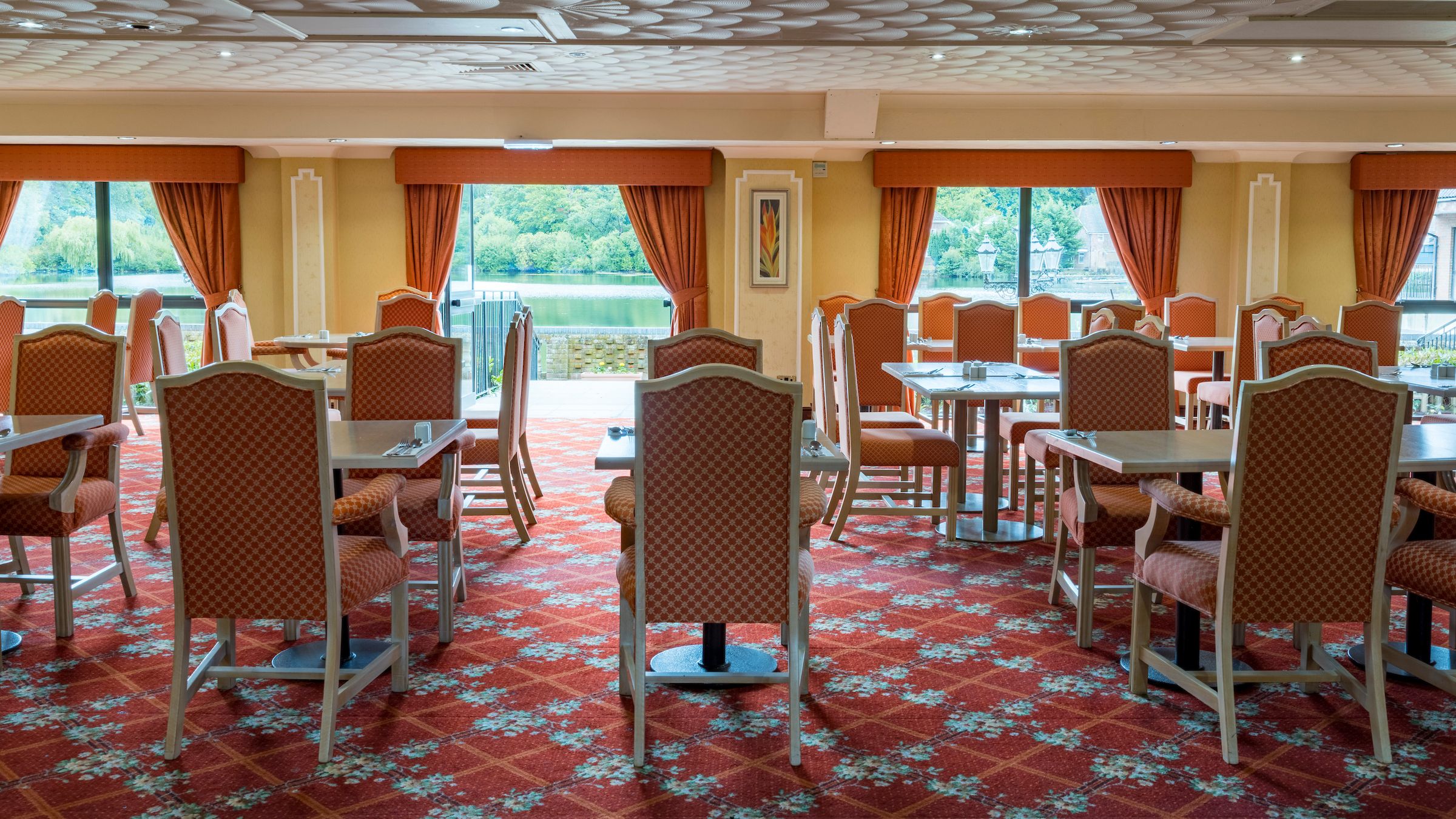 Spacious dining room with red patterned carpet, orange curtains, and large windows overlooking a scenic lake view.