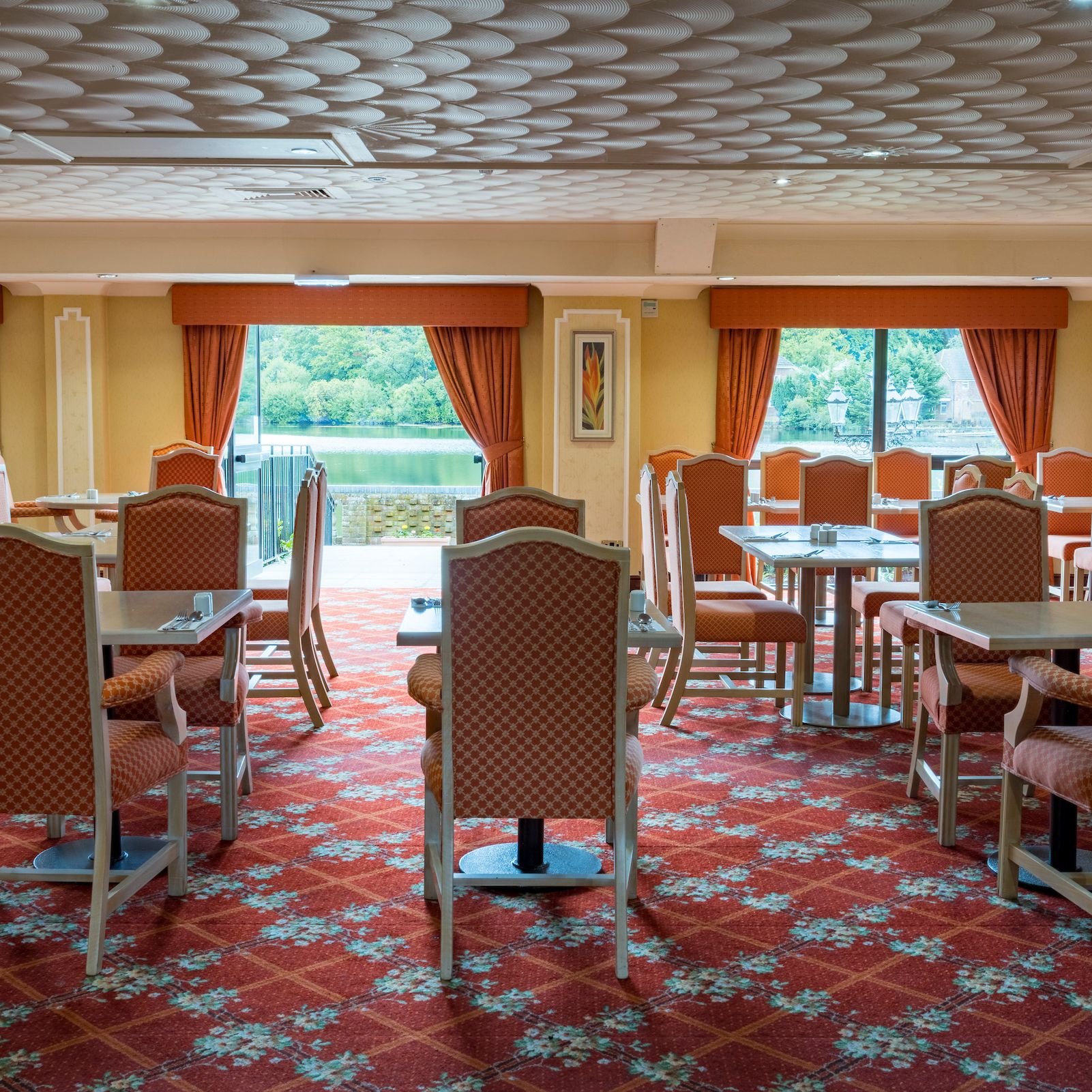 Spacious dining room with red patterned carpet, orange curtains, and large windows overlooking a scenic lake view.