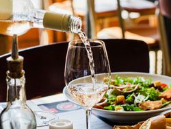 A bottle of white wine being poured into a glass on a restaurant table with salad and bread