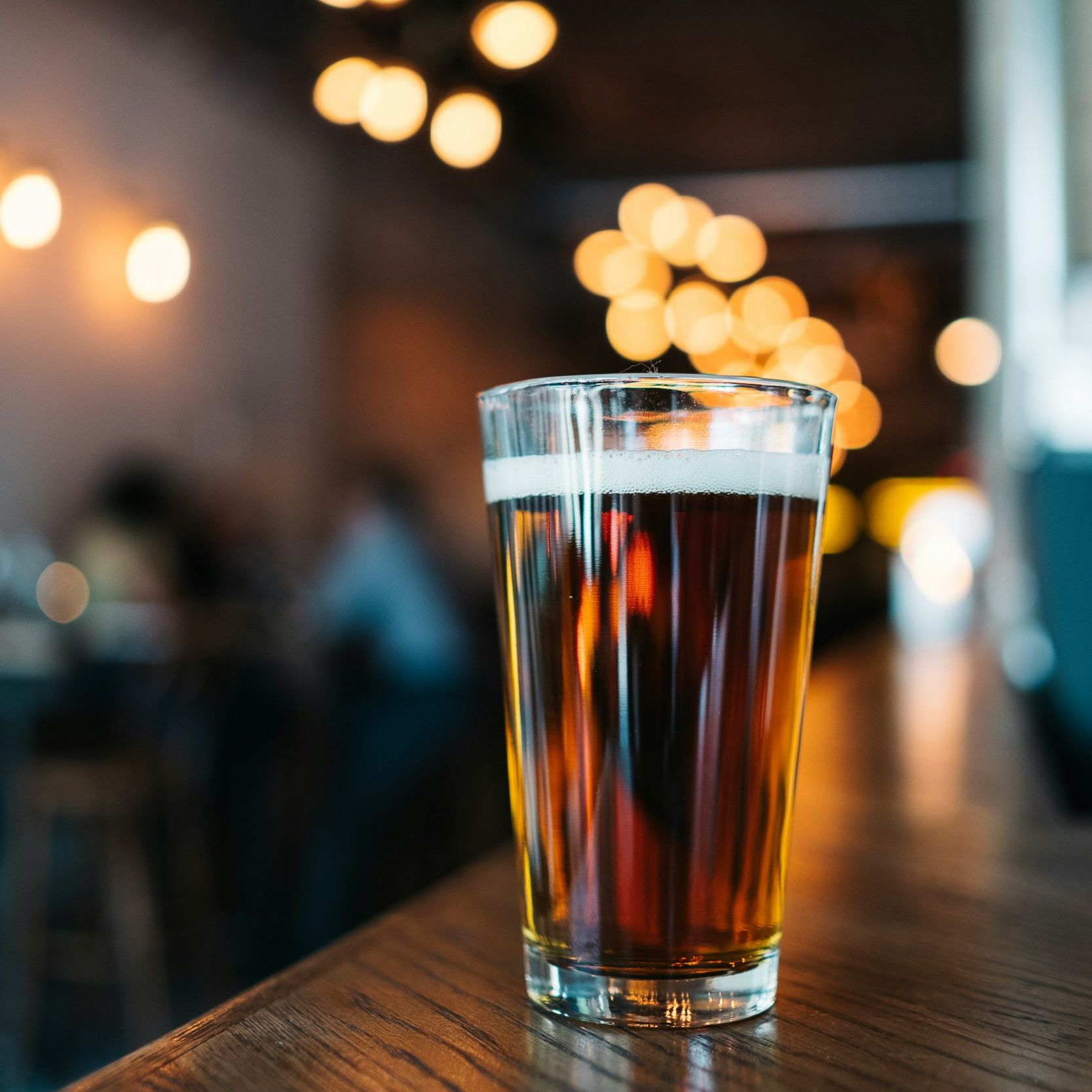 A pint glass filled with amber beer on a wooden bar counter with blurred lights in the background.