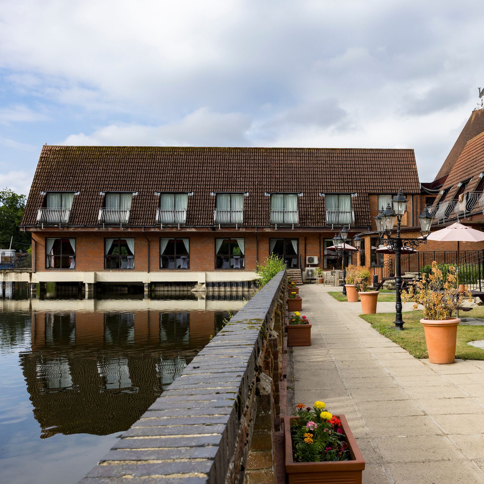 A lakeside hotel with outdoor seating and flower pots along a walkway beside the water.
