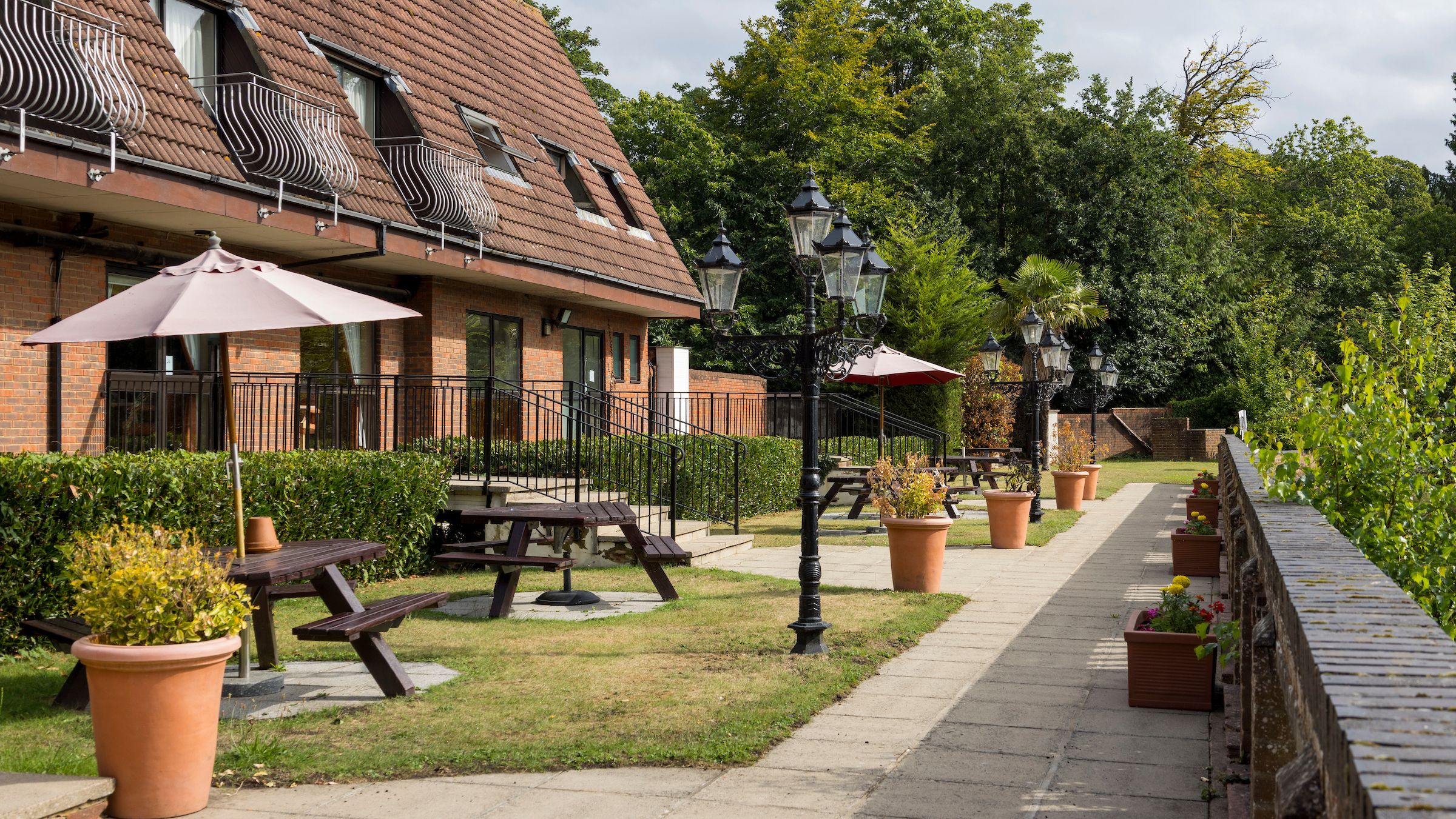 Outdoor seating area with benches, umbrellas, potted plants, and a brick building in the background.