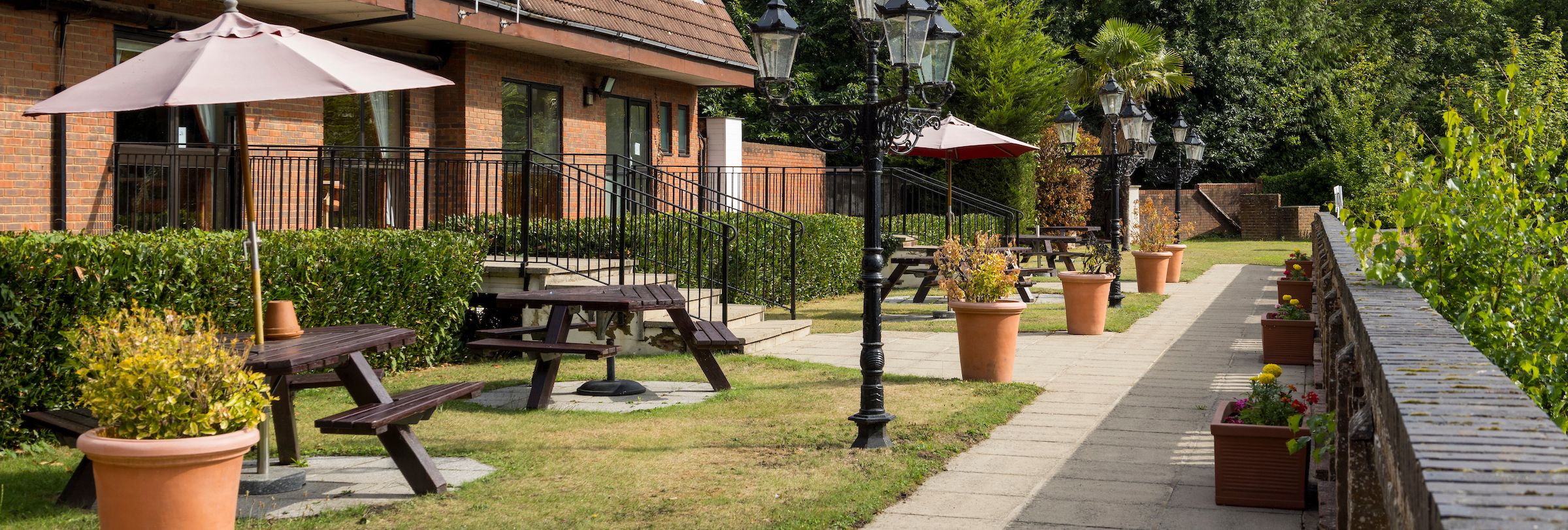 Outdoor seating area with benches, umbrellas, potted plants, and a brick building in the background.