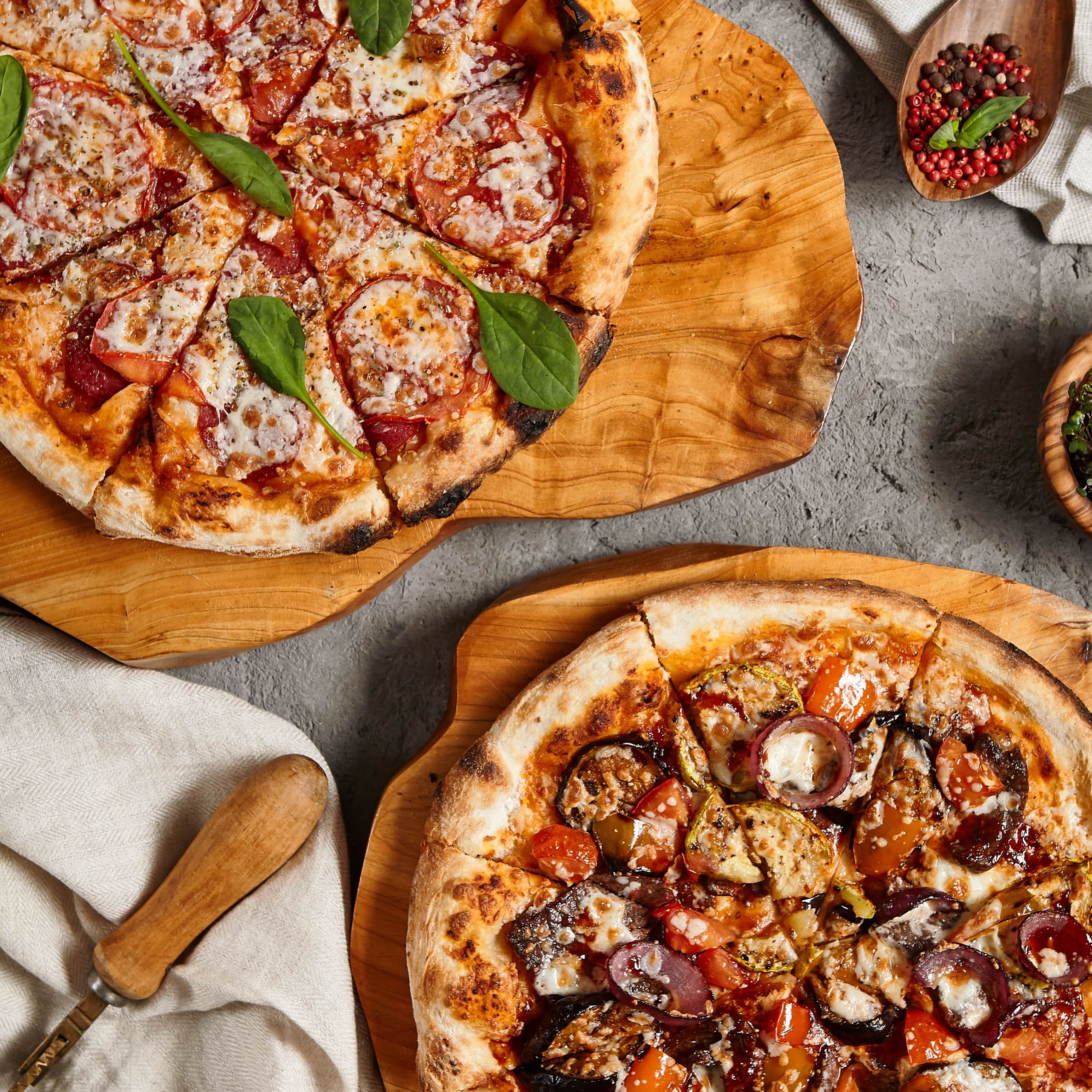 Two gourmet pizzas on wooden boards, one topped with spinach, pepperoni, and cheese, the other with assorted vegetables and cheese.