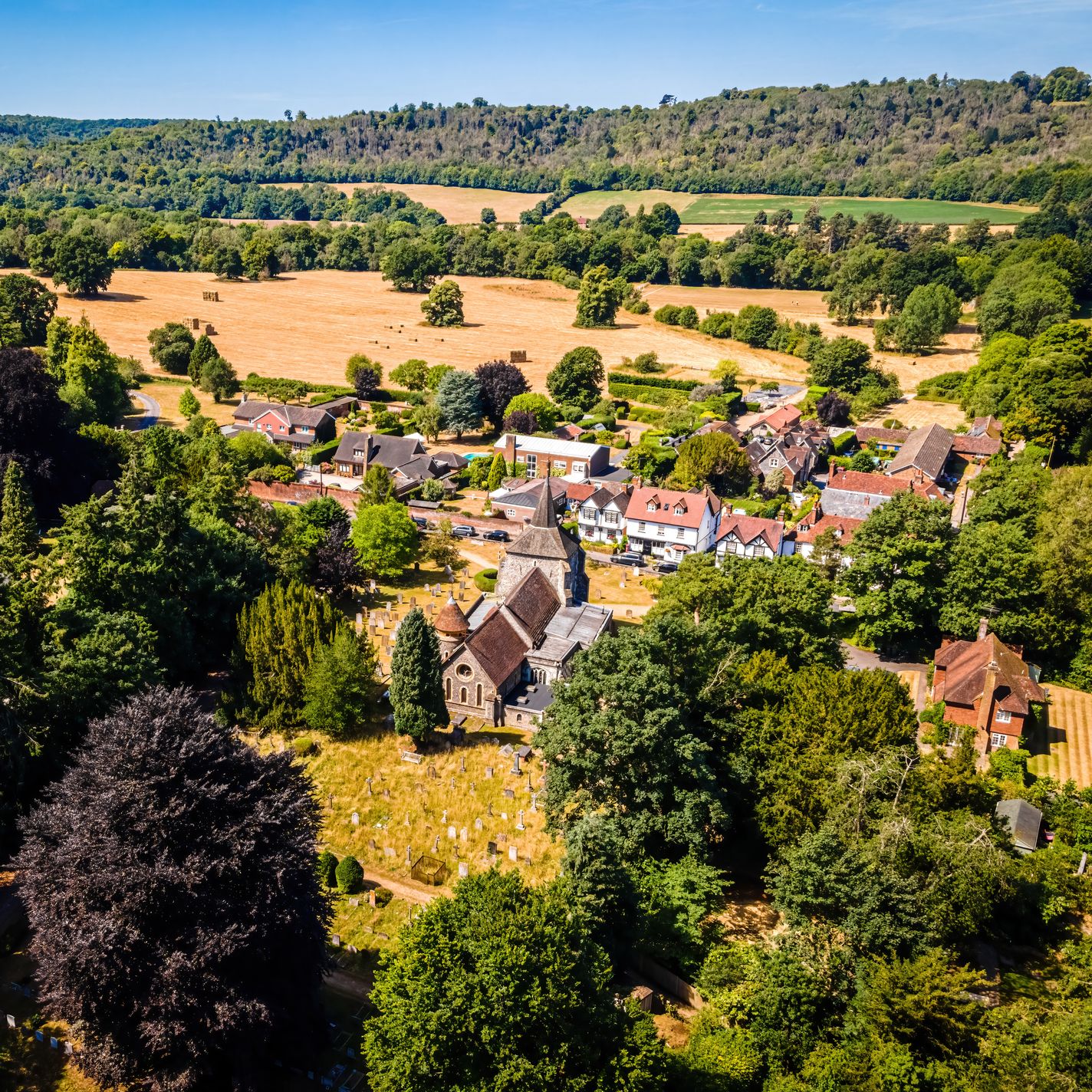 Aerial view of Mickleham, Surrey, showing a village with historic buildings, a church, and surrounding fields and woodland on a sunny day.