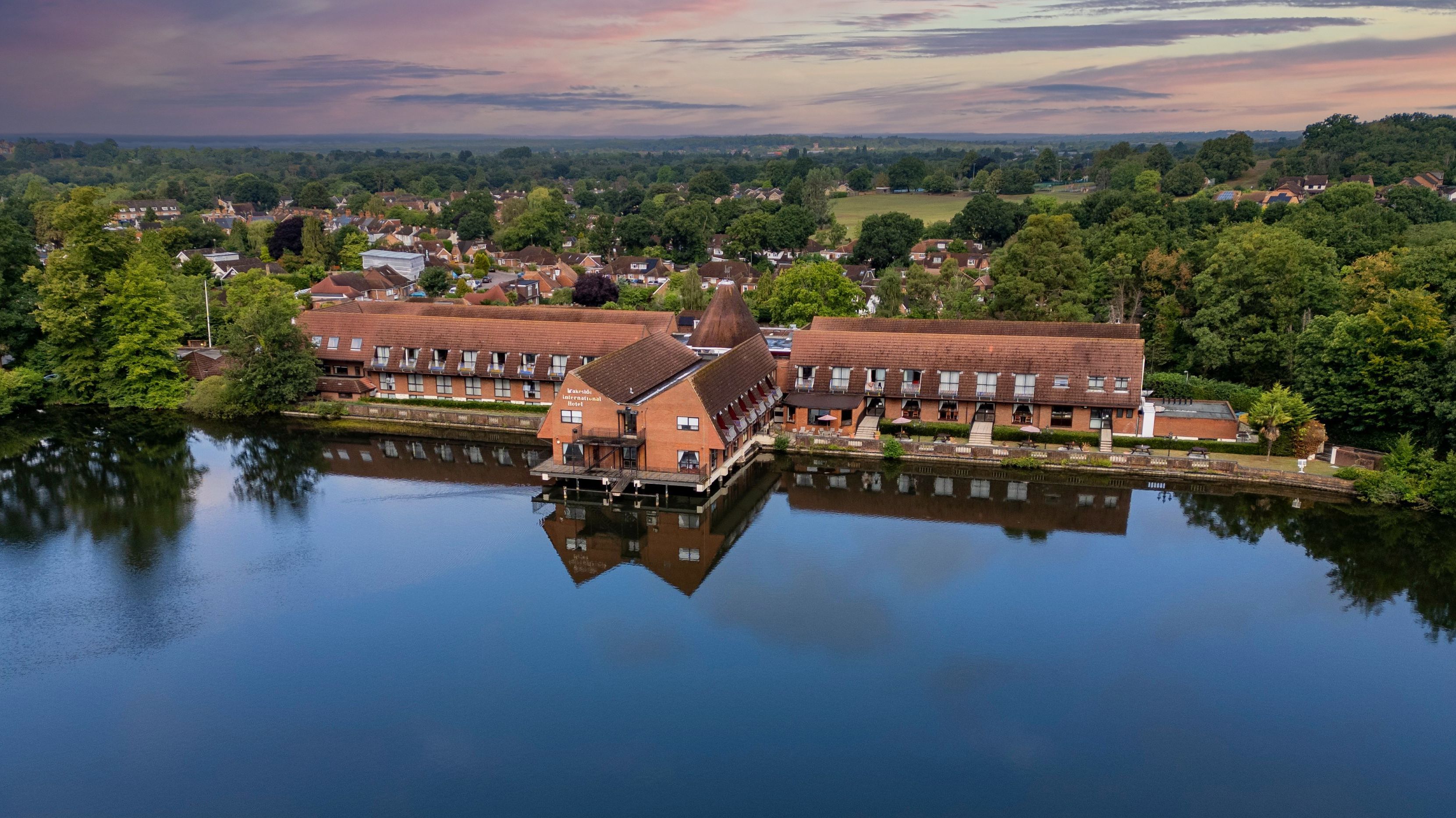 Aerial view of a building complex with red roofs situated on the edge of a calm body of water, surrounded by trees and a residential area in the background during sunset.
