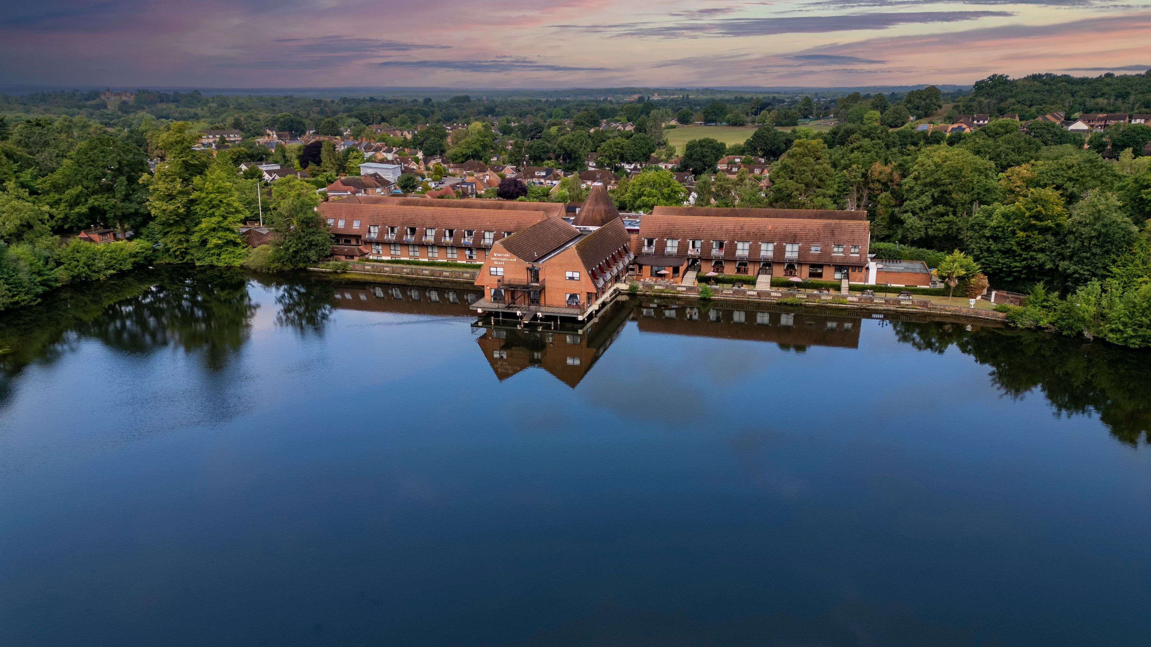 Aerial view of a building complex with red roofs situated on the edge of a calm body of water, surrounded by trees and a residential area in the background during sunset.