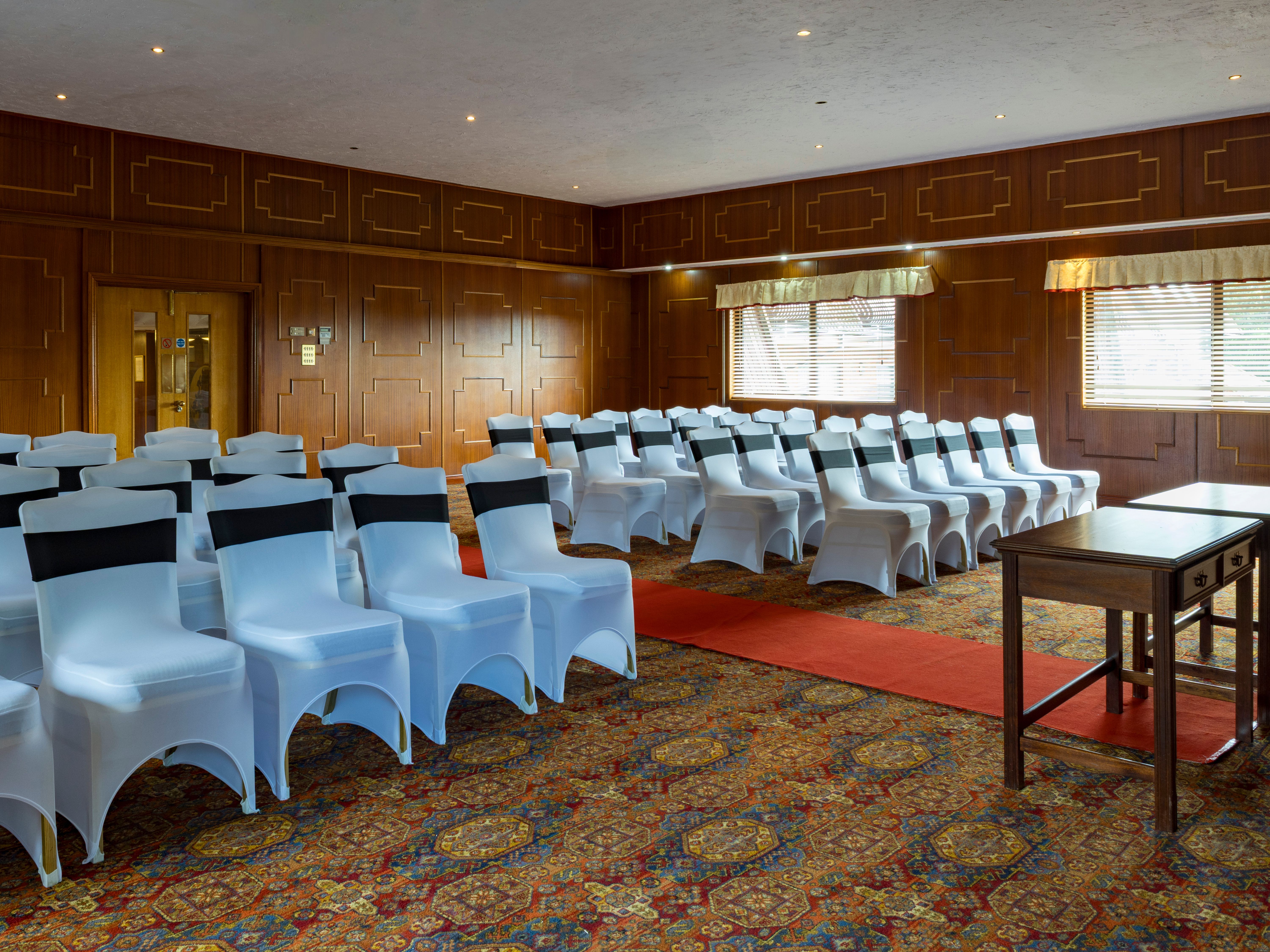 Room set up for an event with white covered chairs, black sashes, red carpet, and wooden tables.