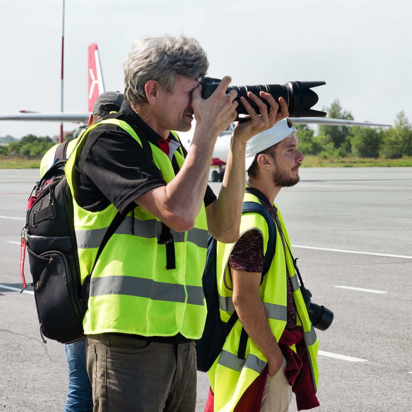 Two men in high-visibility vests taking photographs near an airport runway
