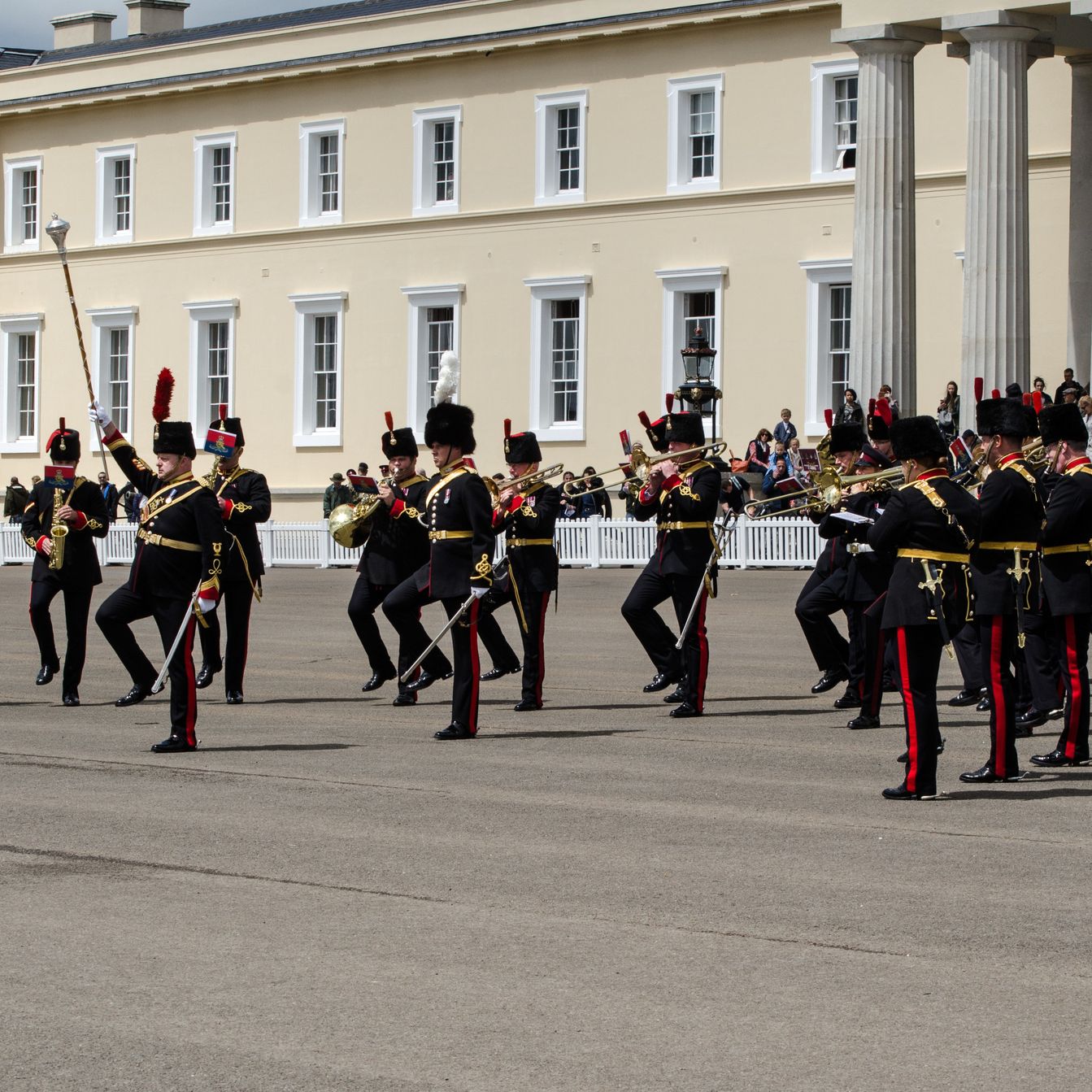 A military band dressed in formal uniforms performs a marching routine in front of a large historic building with columns, while spectators watch in the background.