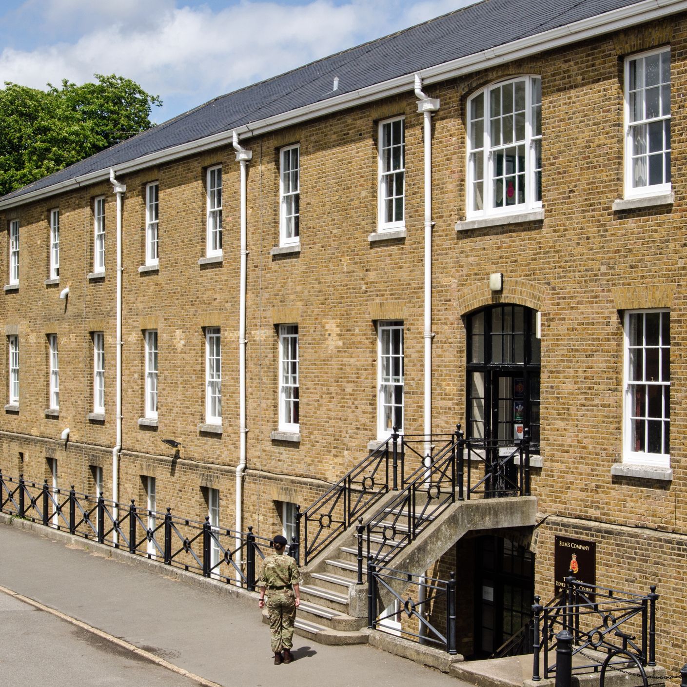 A person in military uniform walks toward the entrance of a large brick building with white-framed windows and black railings. Trees and a blue sky with clouds are in the background.