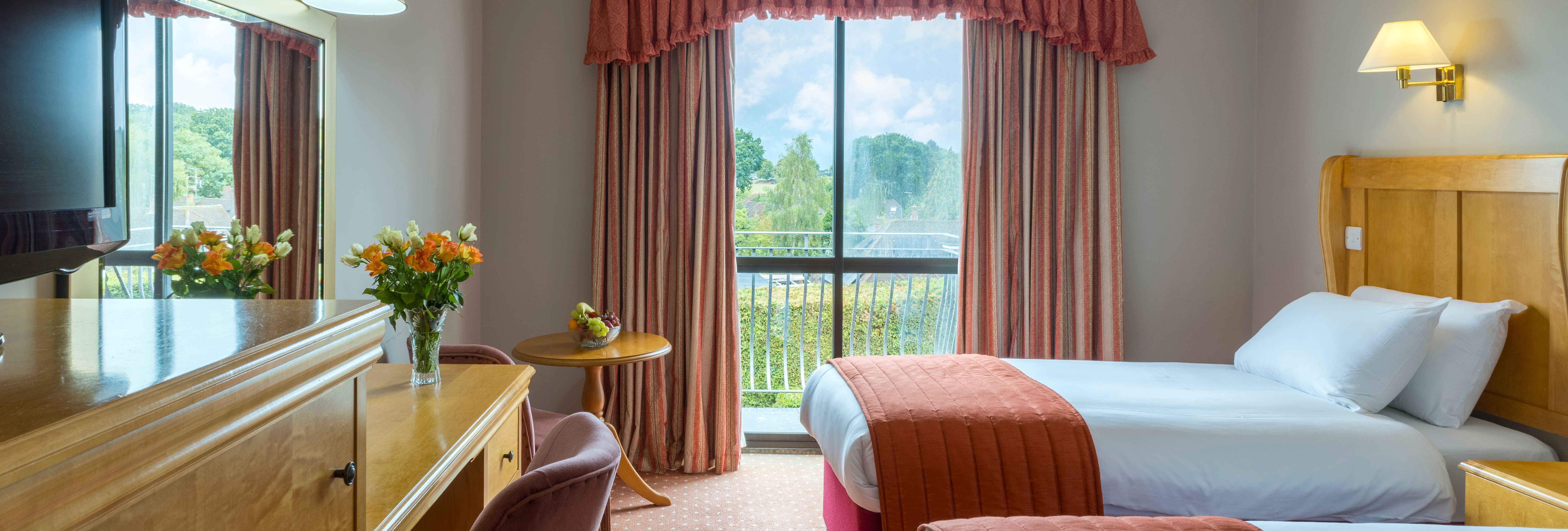 A bright hotel room with two beds, a desk, flowers, and a large window with drapes.